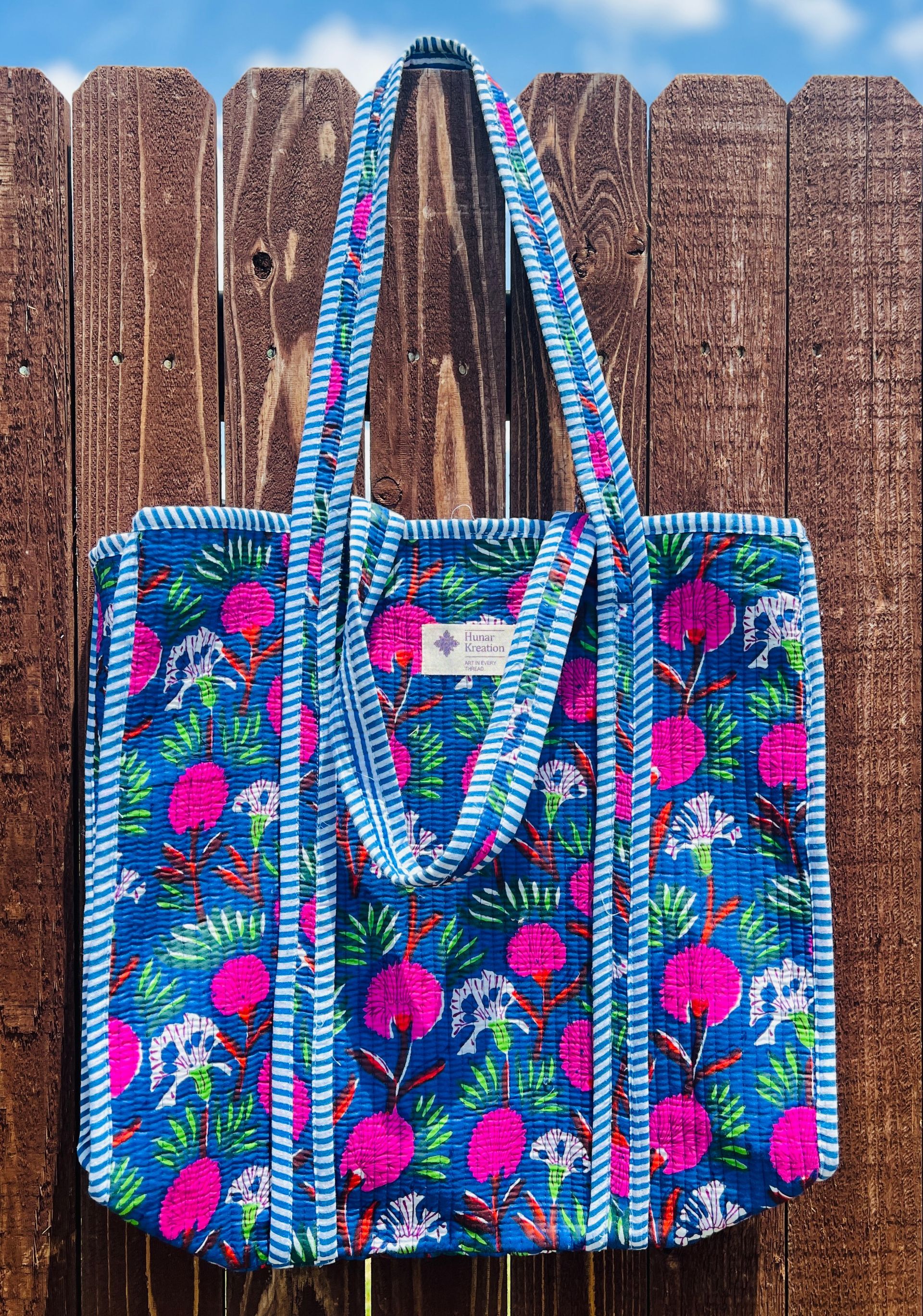 A blue tote bag with pink flowers on it is hanging on a wooden fence.