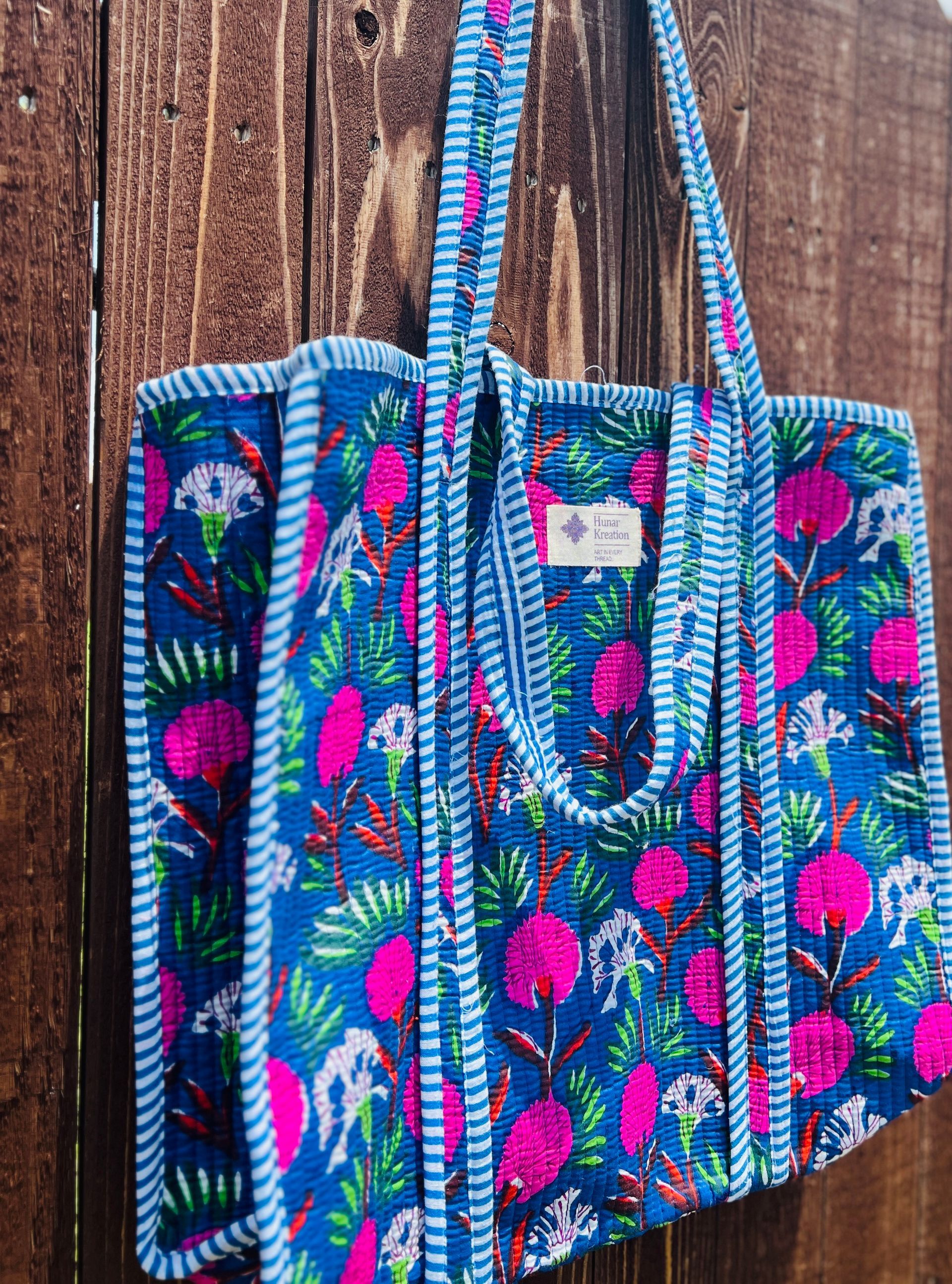 A blue tote bag with pink flowers on it is hanging on a wooden fence.