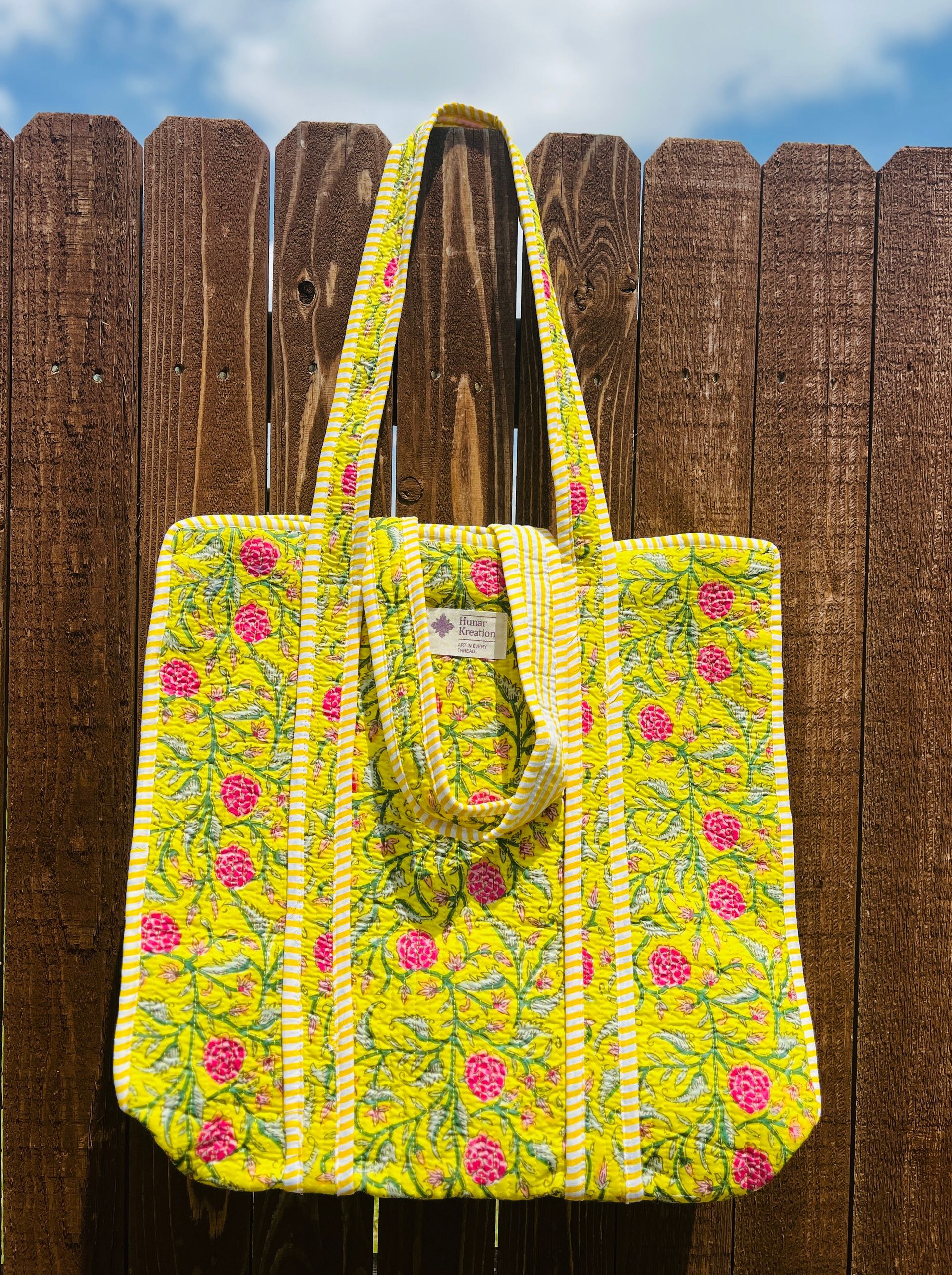 A yellow tote bag with pink flowers is hanging on a wooden fence.