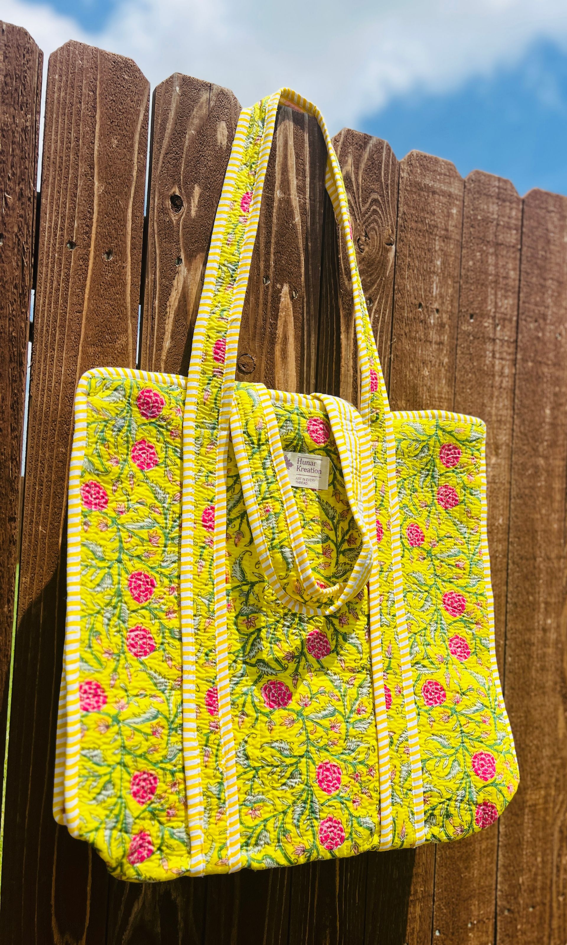 A yellow tote bag with pink flowers is hanging on a wooden fence.