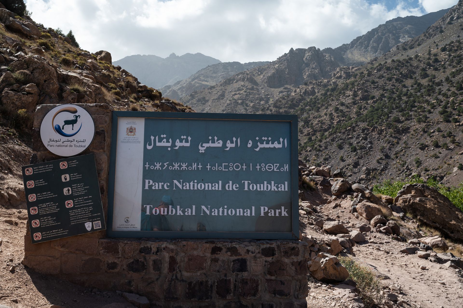 A rectangular sign in Toubkal National Park, Morocco, set against a backdrop of rugged, rocky mountains.