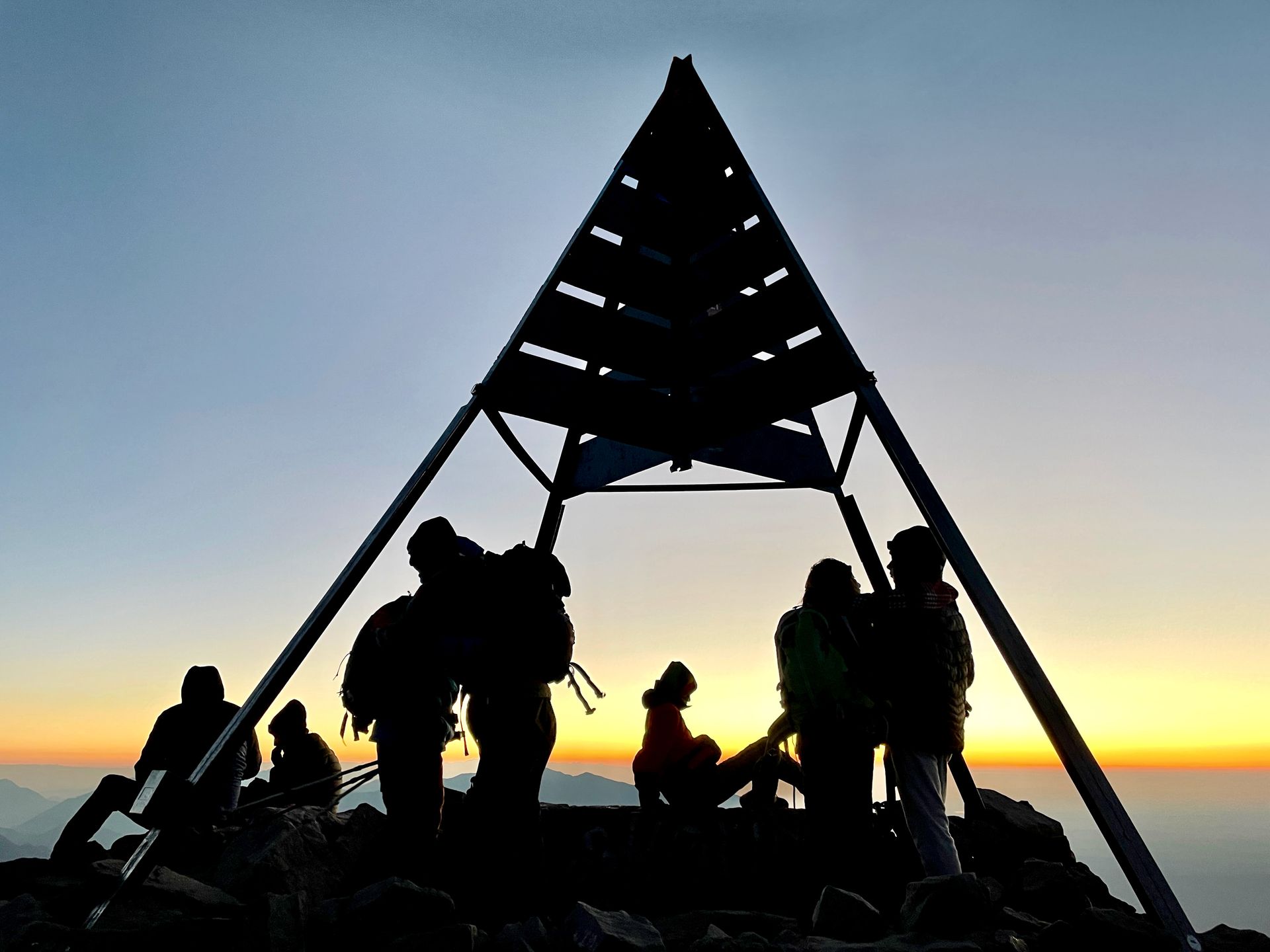 Silhouetted hikers stand beneath a triangular metal summit marker against a vibrant sunrise over mountain peaks.