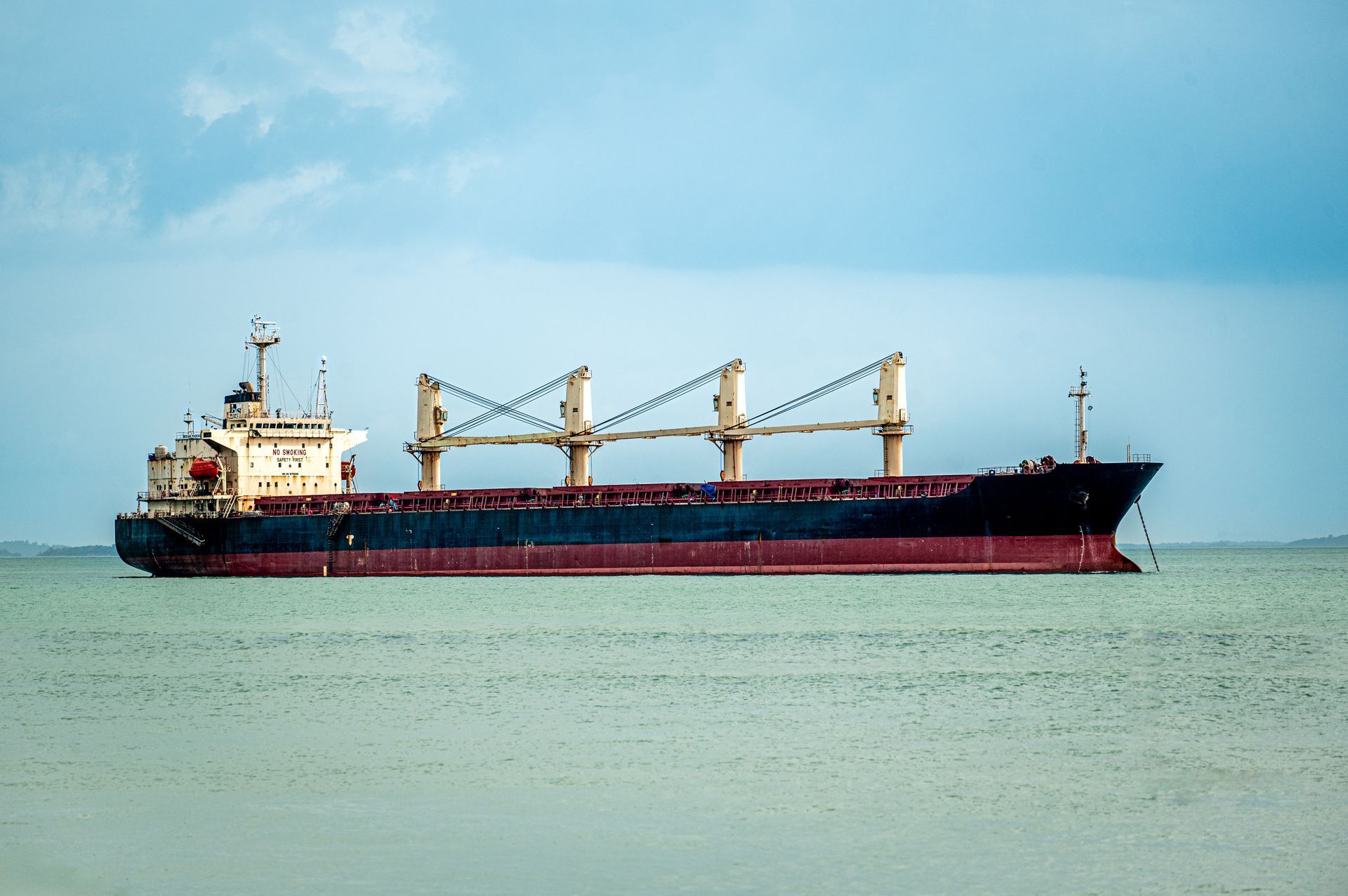 A large, dark-hulled cargo ship with four cranes on its deck sits on calm, light-colored water under a cloudy blue sky.