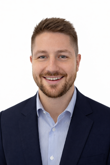Headshot of a person with a beard, smiling, wearing a light blue striped collared shirt and a dark navy blazer.