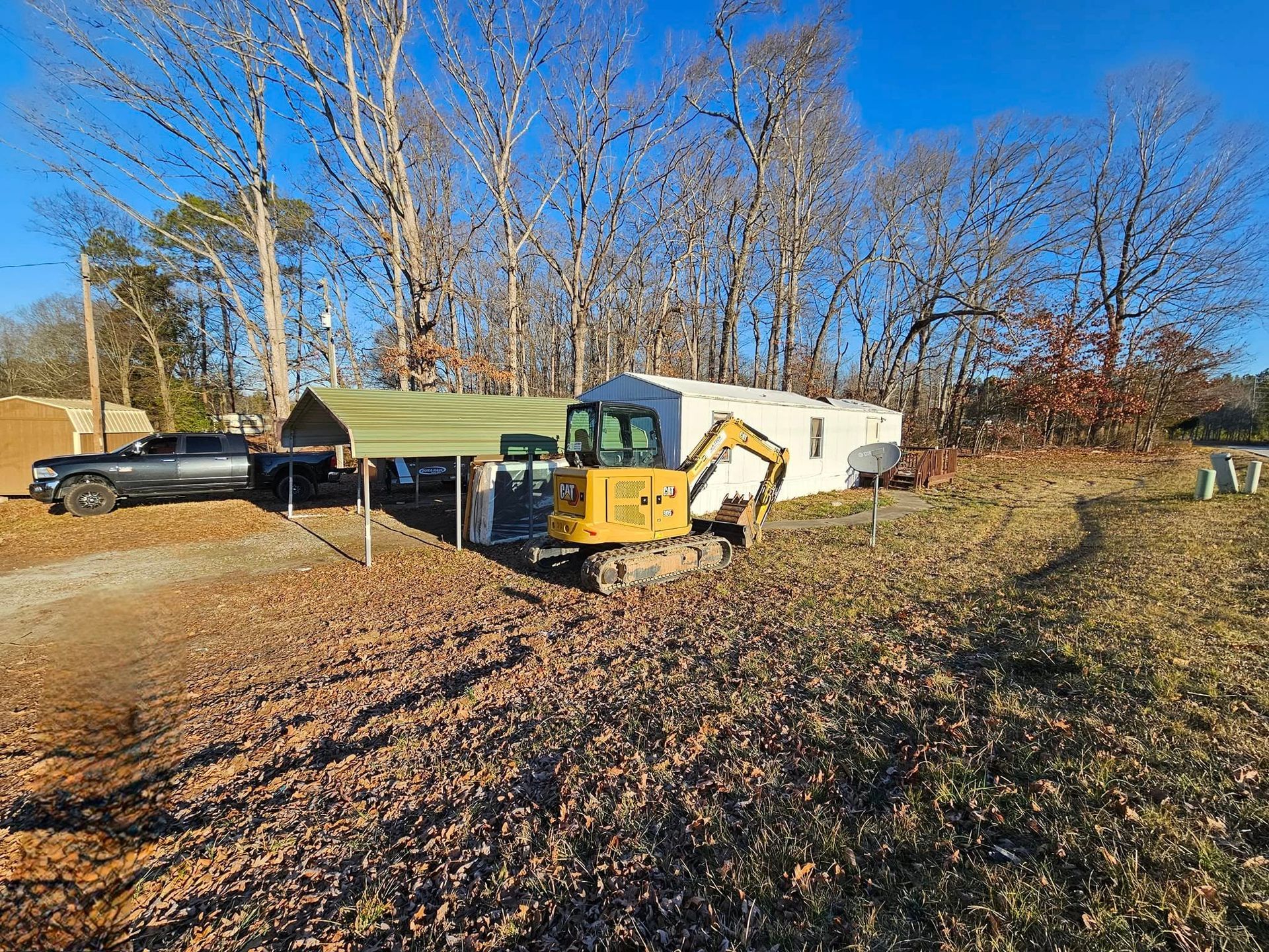 Yellow excavator parked near white shed under a tarp, on a fall-colored field, with a truck.
