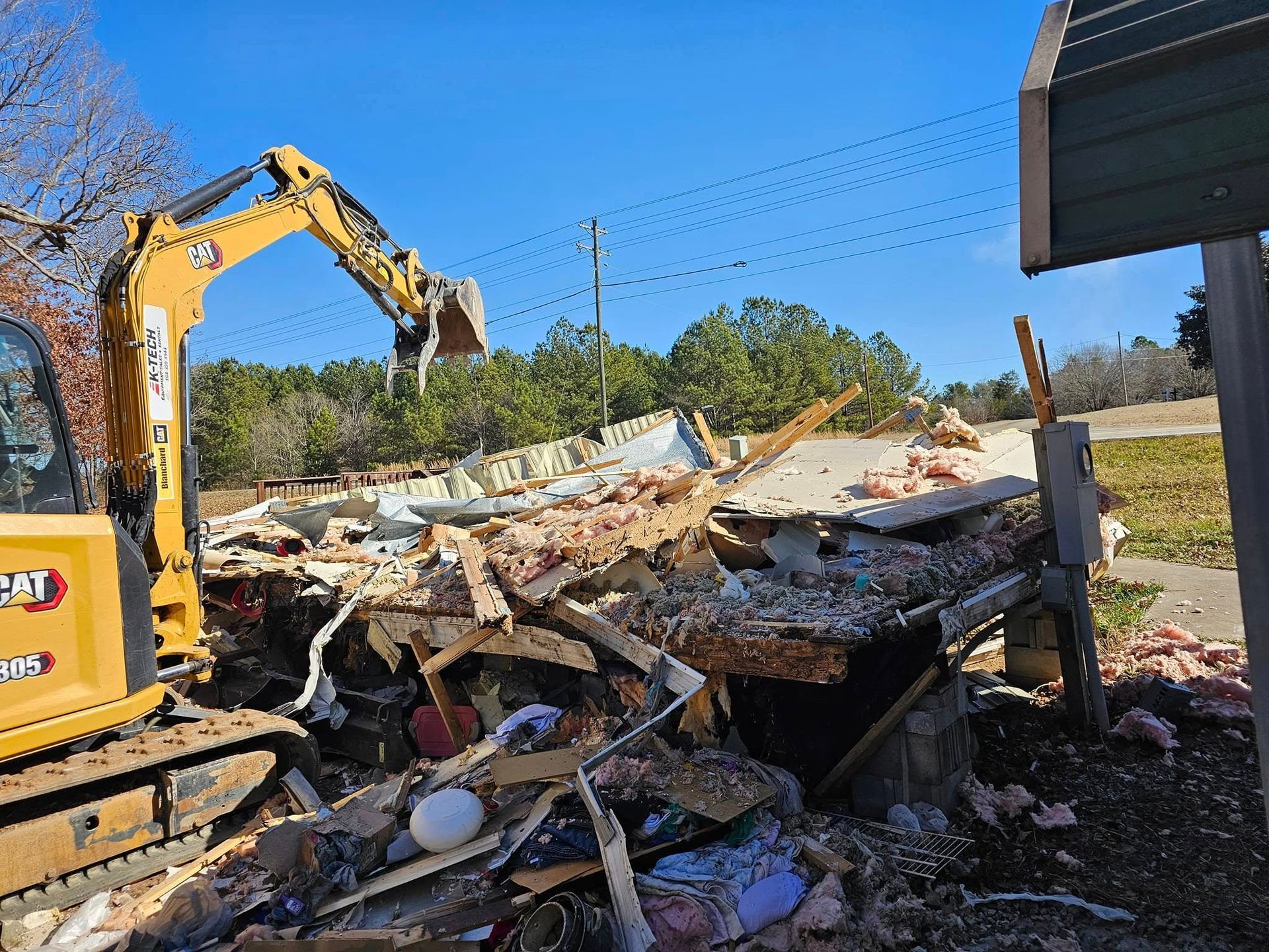 A yellow excavator demolishes a building on a sunny day; debris and wooden supports are scattered.
