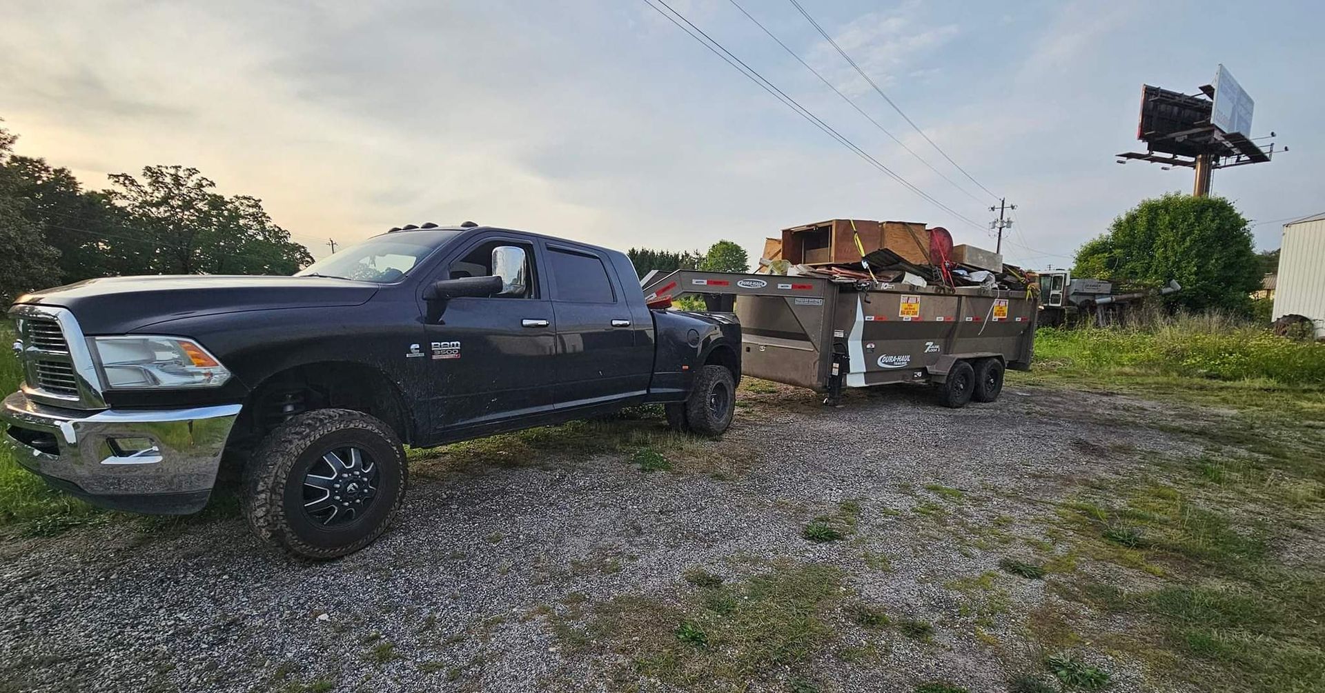 Black truck towing a trailer loaded with scrap metal on a gravel surface.