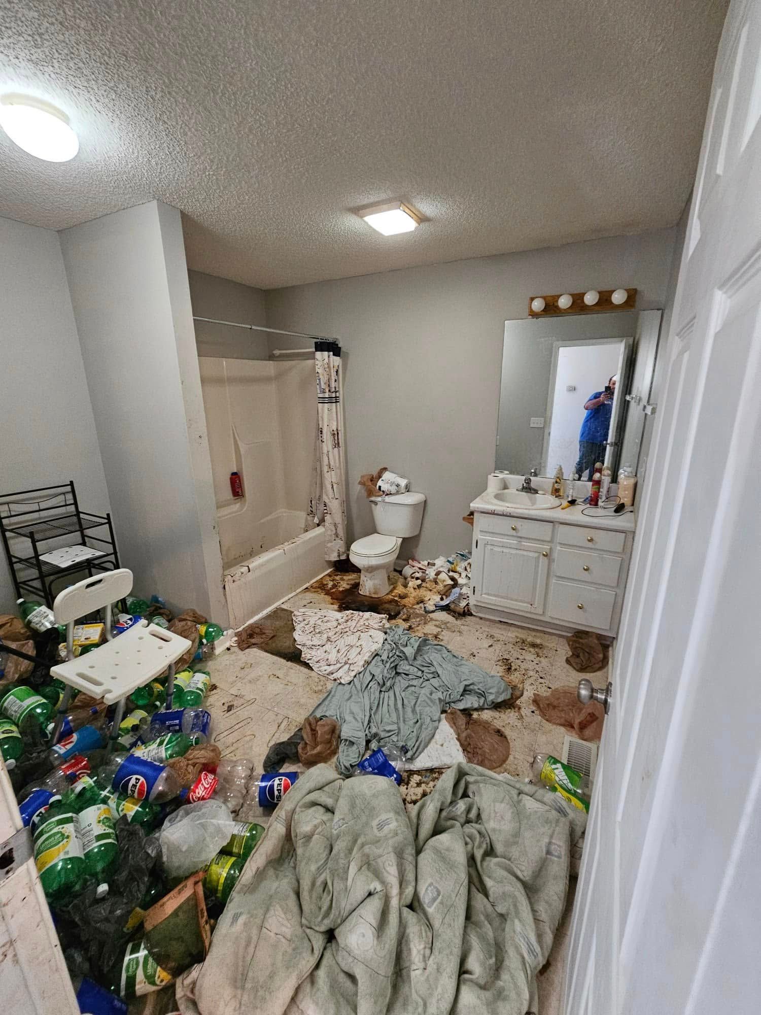 Bathroom in disarray, covered in trash, clothing, and debris.