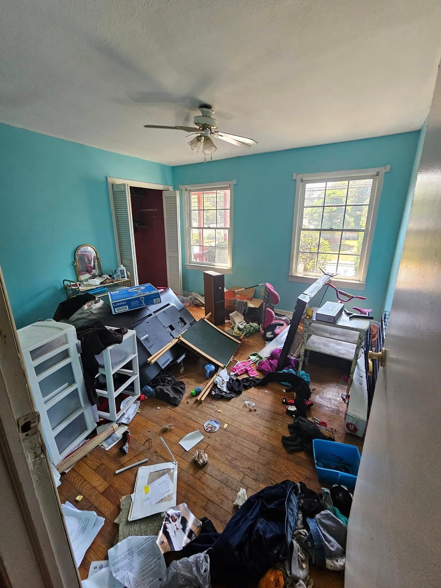Messy bedroom with blue walls, hardwood floor, and scattered items; a ceiling fan and two windows visible.