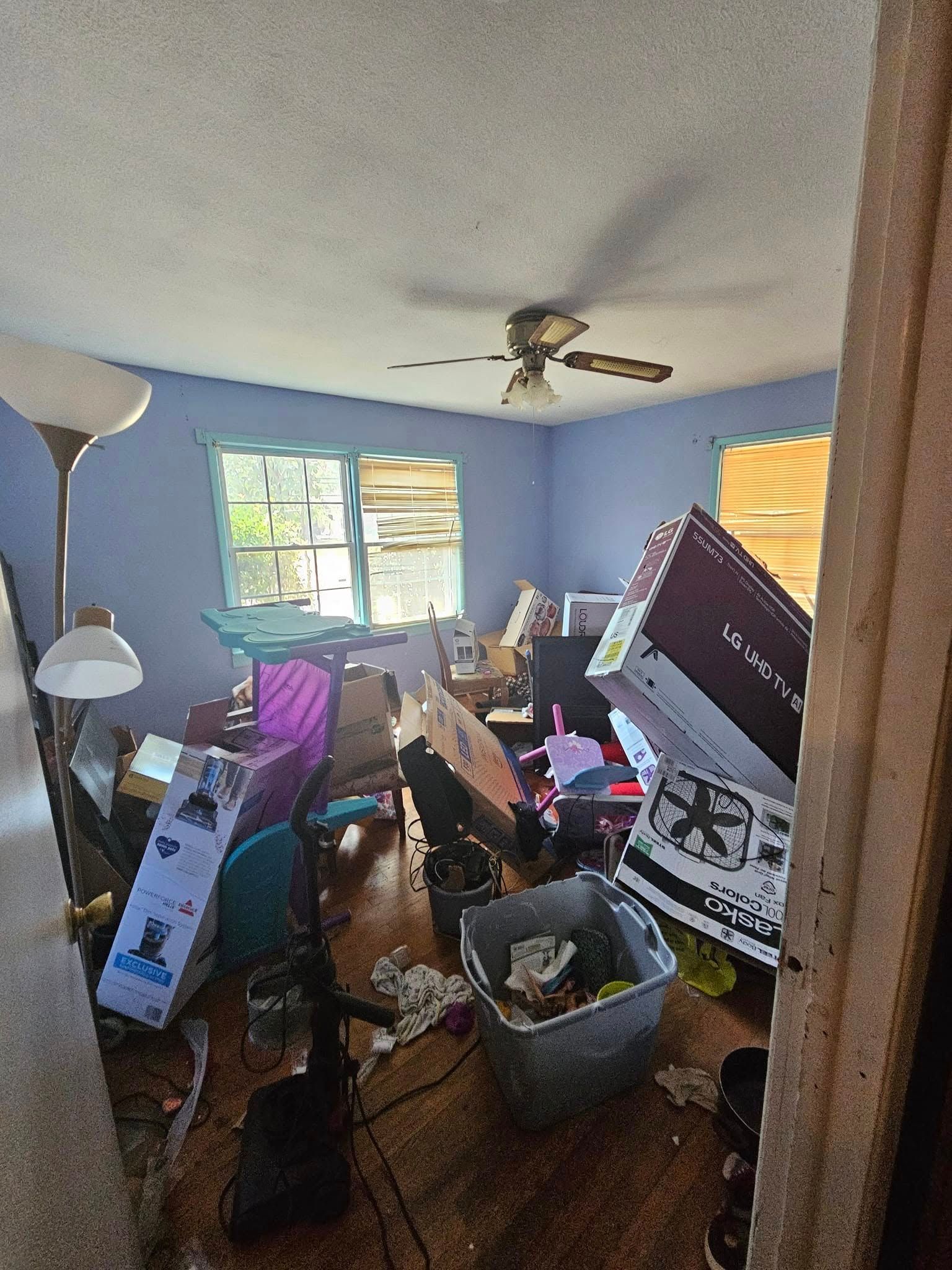 Messy bedroom with boxes, trash, and debris scattered on the floor. Blue walls, window, ceiling fan visible.