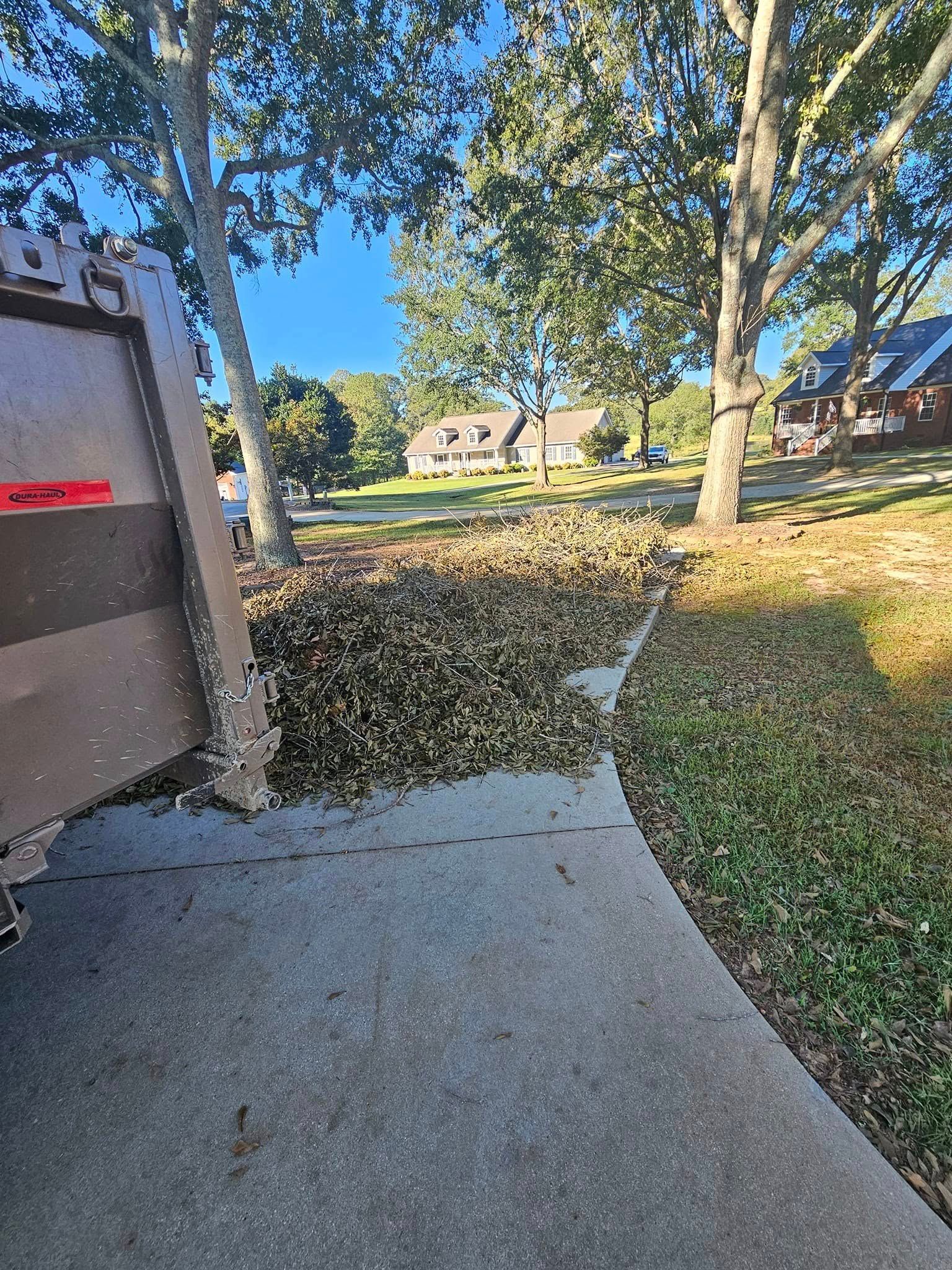 A brown garbage truck dumping leaves onto a driveway, with trees and houses in the background.