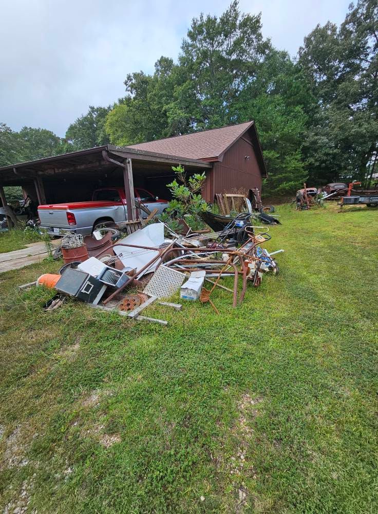 Pile of scrap metal on grass in front of a shed and a covered car parking area with a red truck.