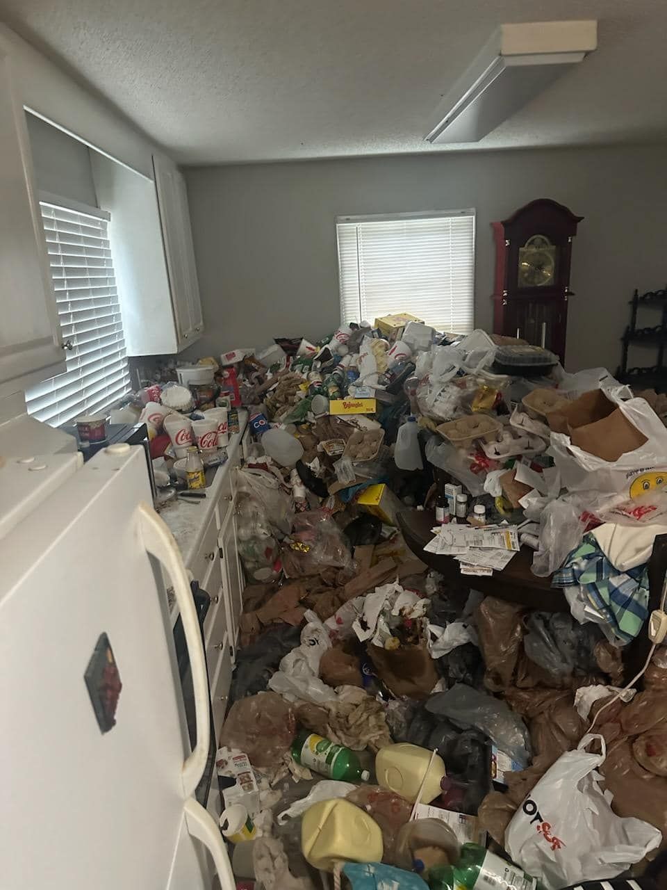 Room filled with a large pile of trash; a refrigerator and clock are visible in the cluttered space.