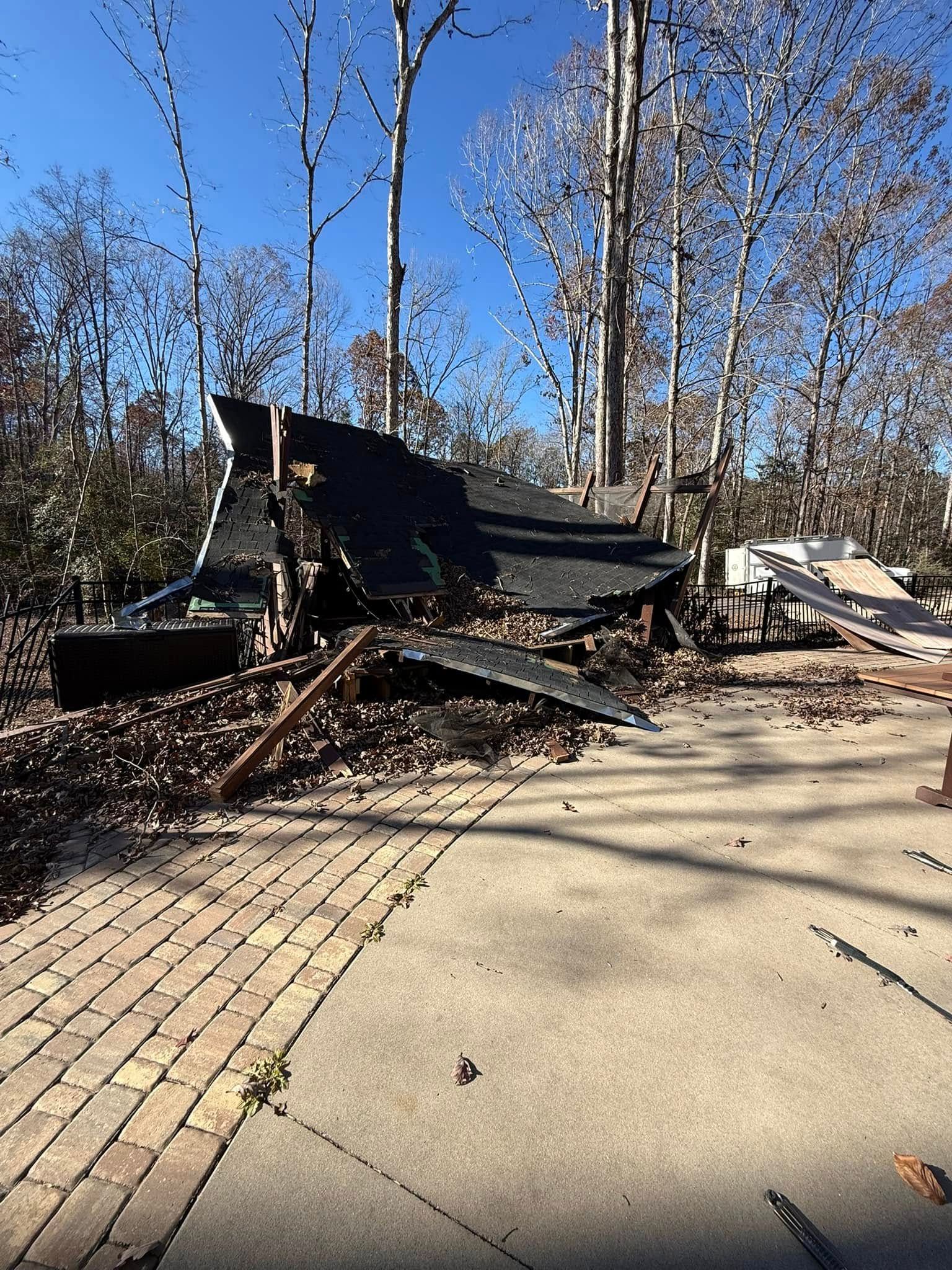 A yellow excavator demolishes a building, tearing away roof and walls. Construction worker on left.