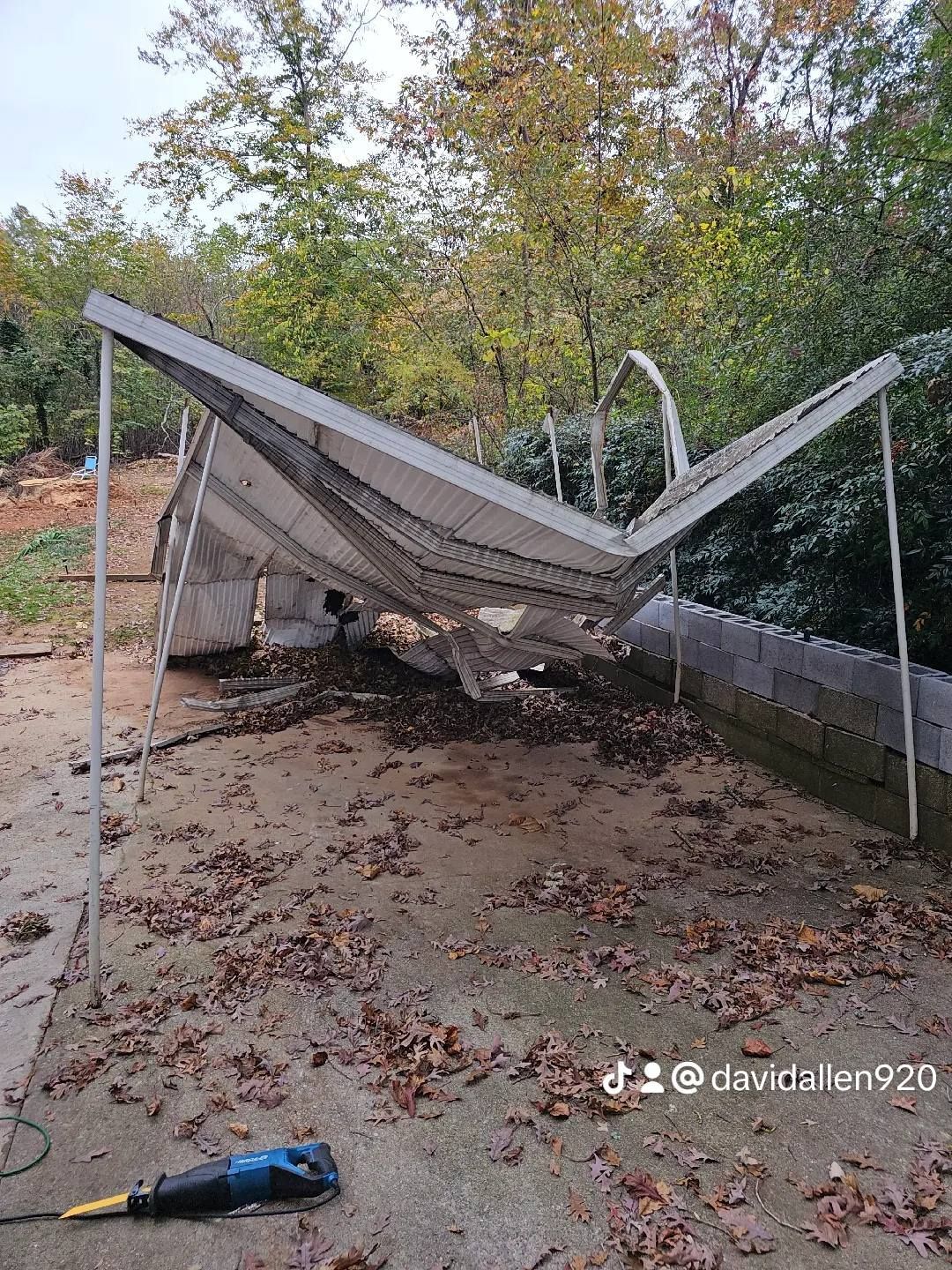 Collapsed white canopy structure with fallen leaves, set against trees and a concrete wall.