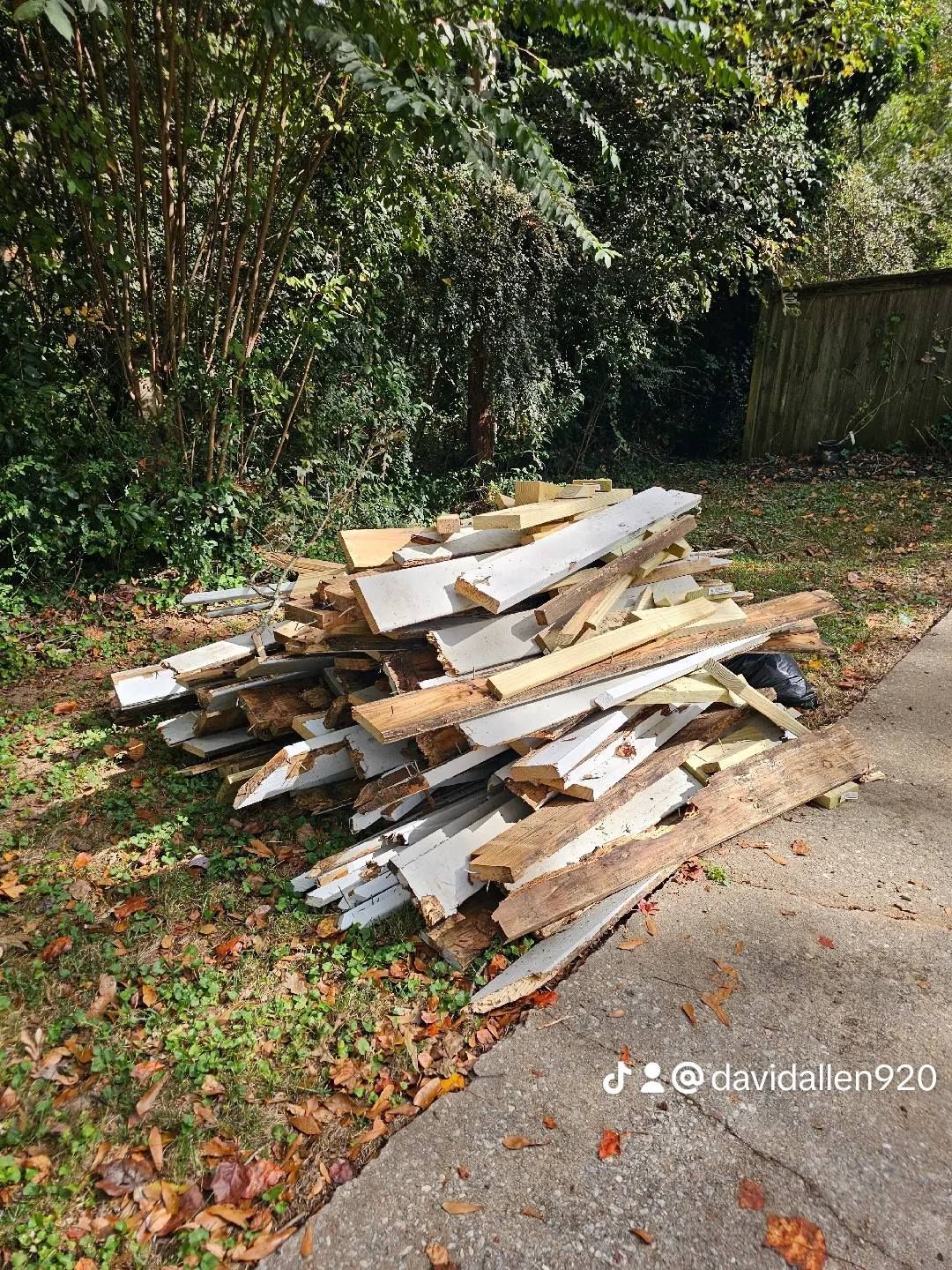 Pile of weathered wood on grass beside a paved surface; trees in the background.
