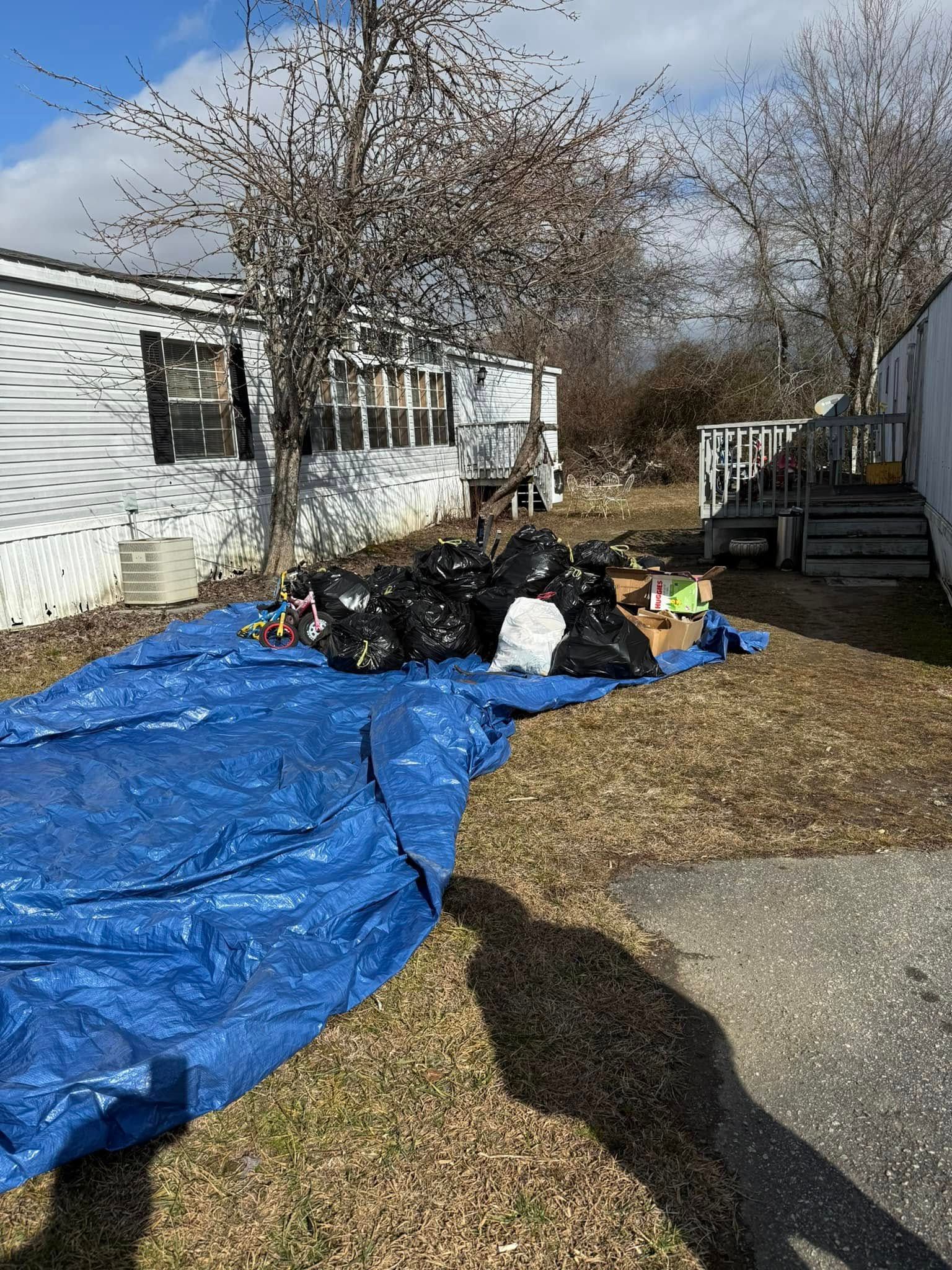 Black trash bags and debris on a blue tarp in a yard near a white mobile home.