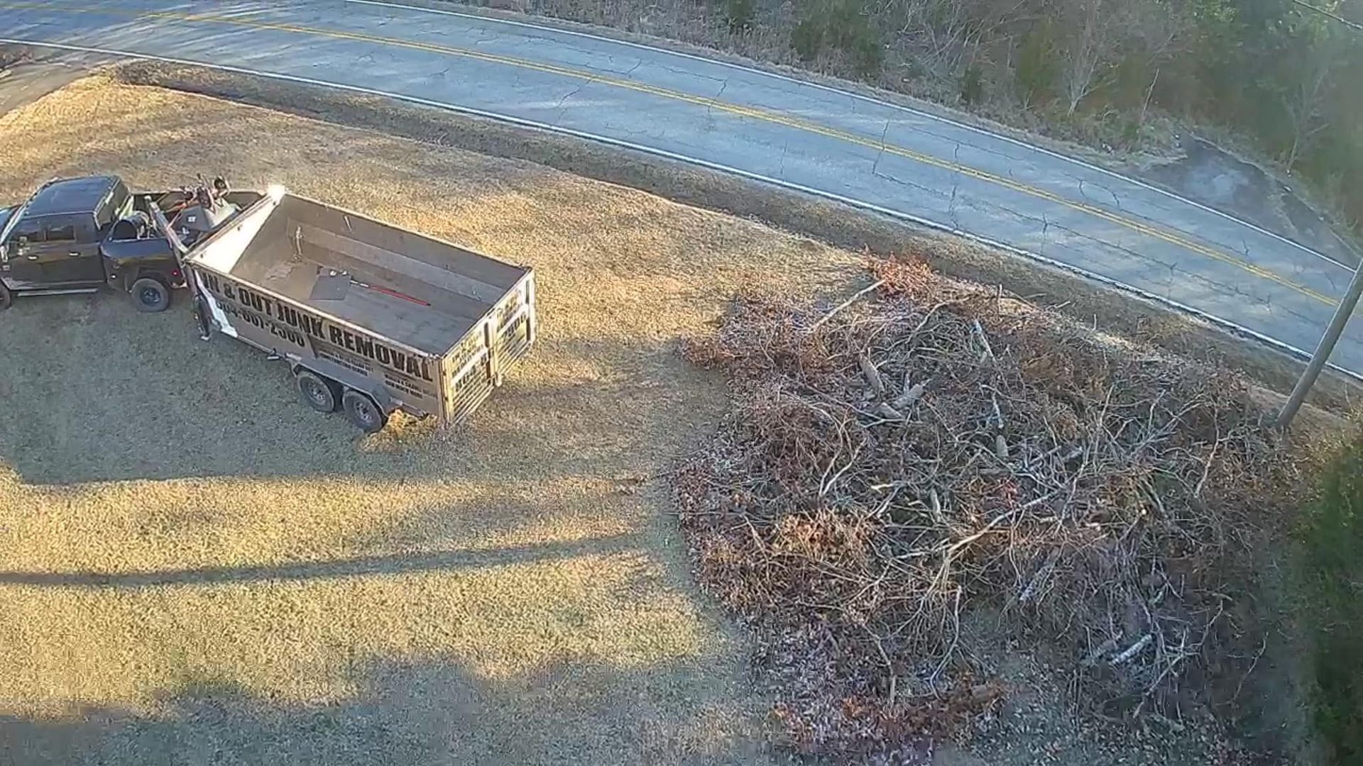 Truck with trailer next to a pile of branches, parked on a grassy field near a road.