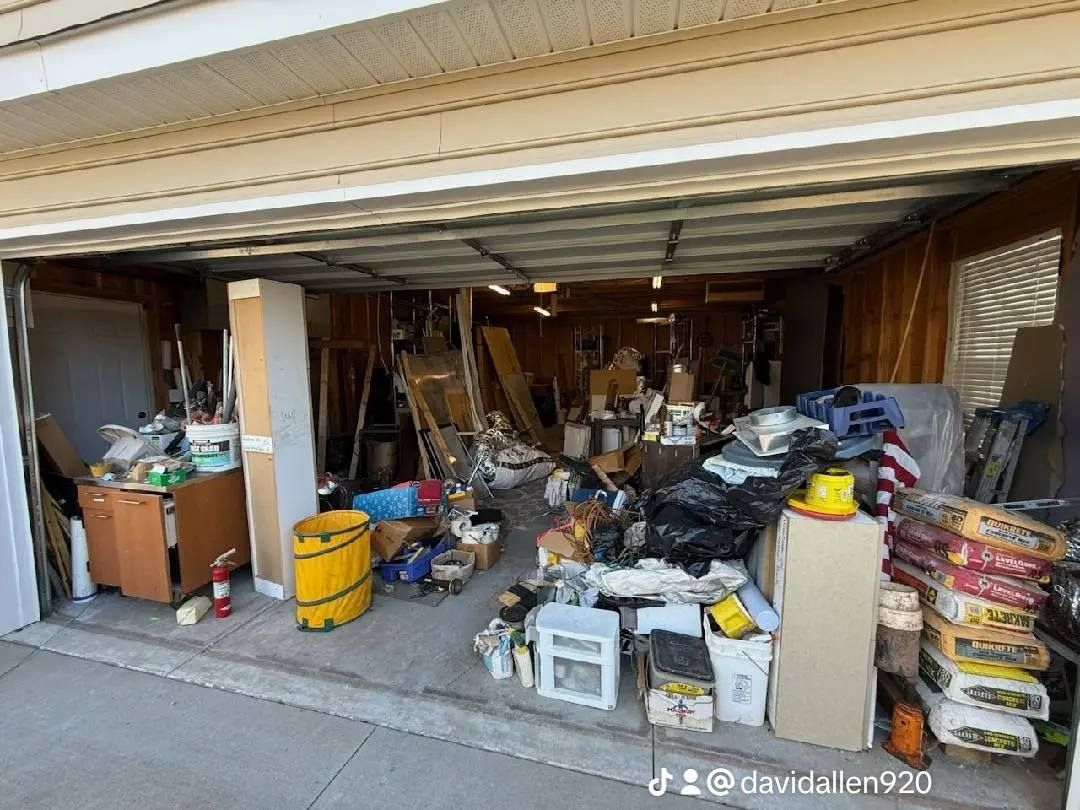 Cluttered garage with various items: boxes, tools, and trash bags. Beige garage door open.