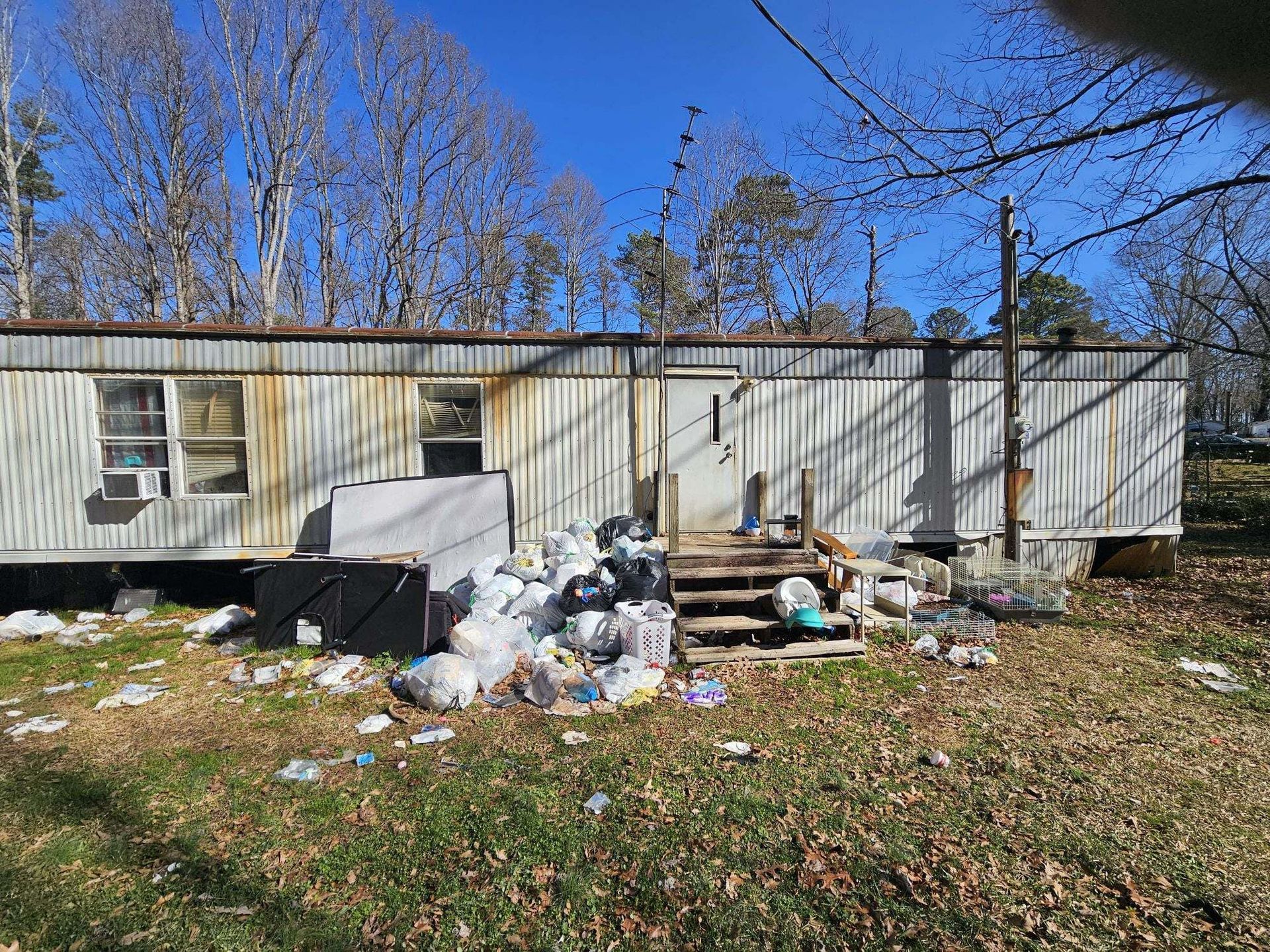Dilapidated trailer surrounded by piles of trash on a brown grassy lawn under a blue sky.