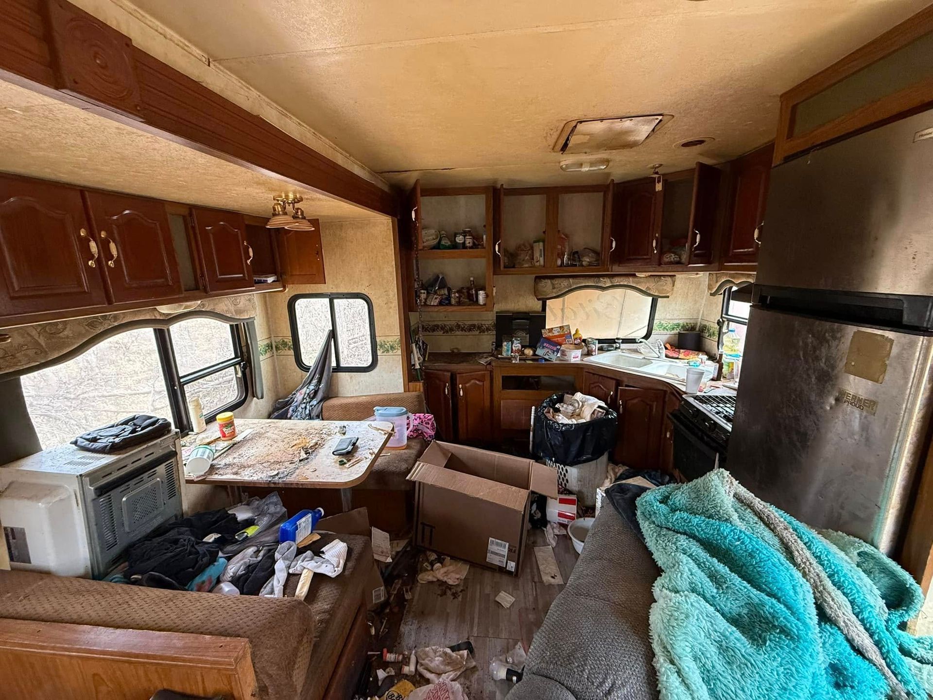 Messy RV interior with debris-covered table, overflowing trash, and disorganized kitchen area.