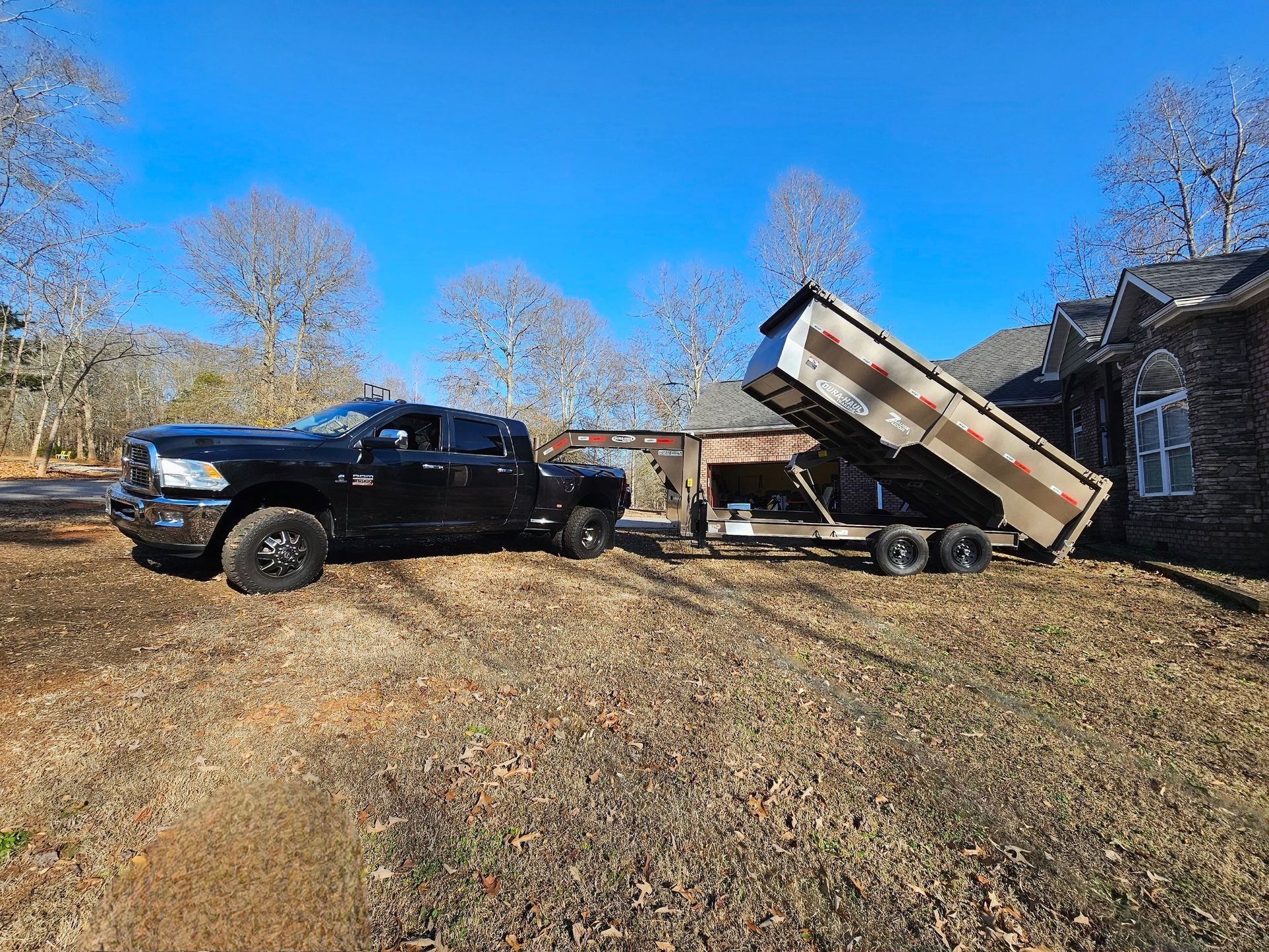 Black pickup truck towing a dump trailer in a yard on a sunny day. Trailer bed is raised.
