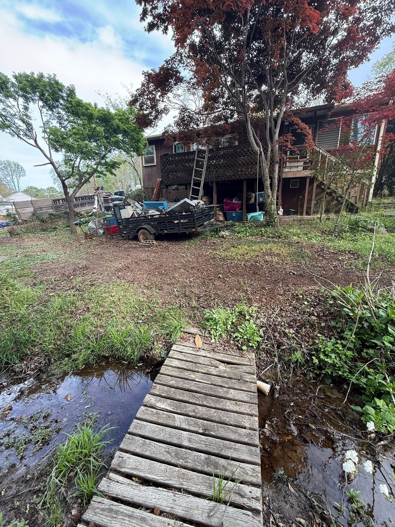 Wooden bridge leads to a house with a brown brick exterior, overgrown vegetation surrounds.