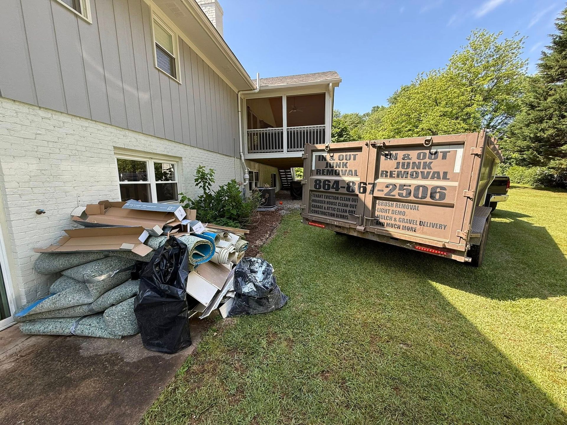 Dumpster next to a house with trash, including cardboard and bags, on the lawn. Brown dumpster.