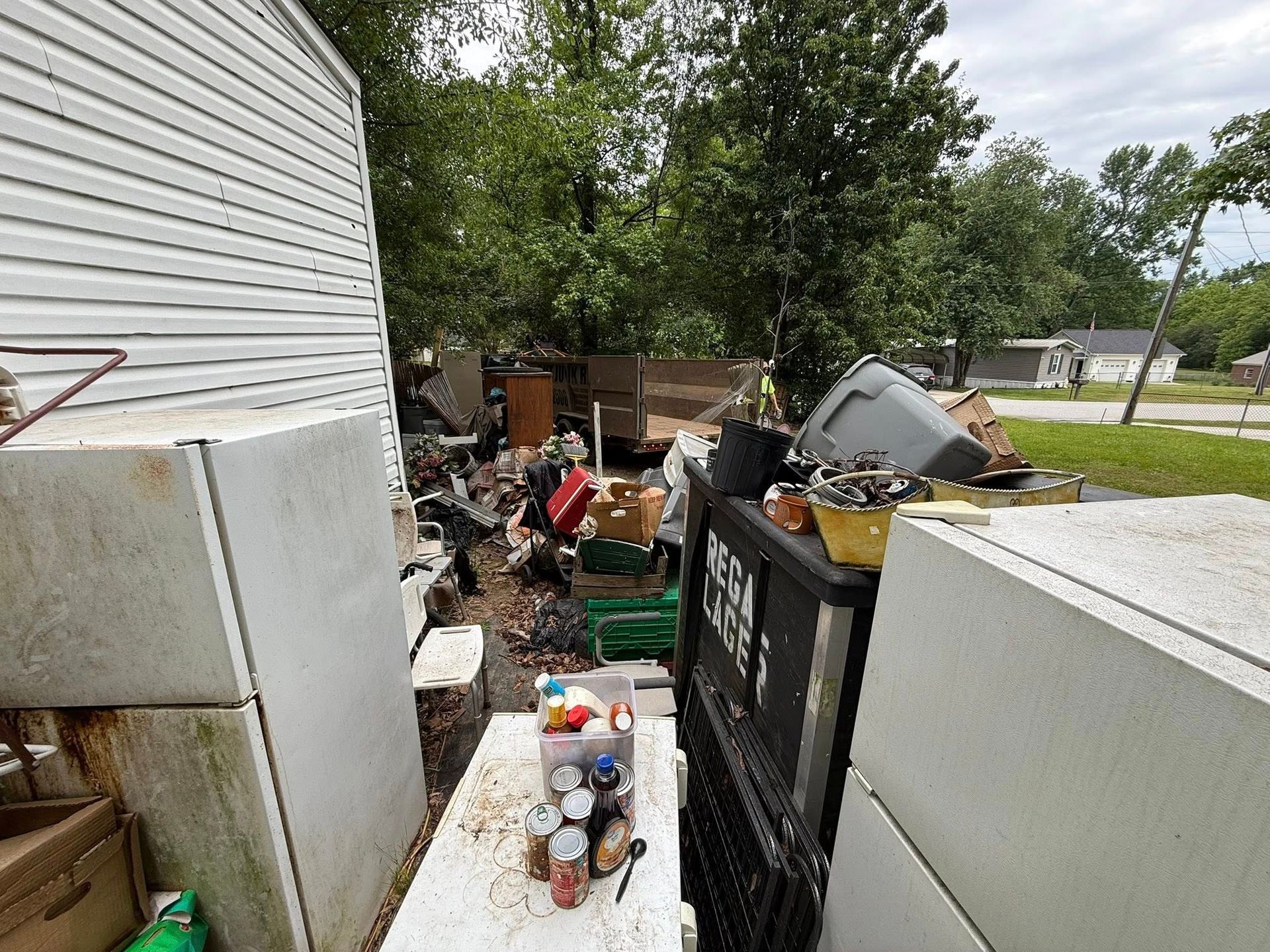 Yard debris: fridge, trash, boxes, and food in a cluttered yard. Trees and a house in the background.