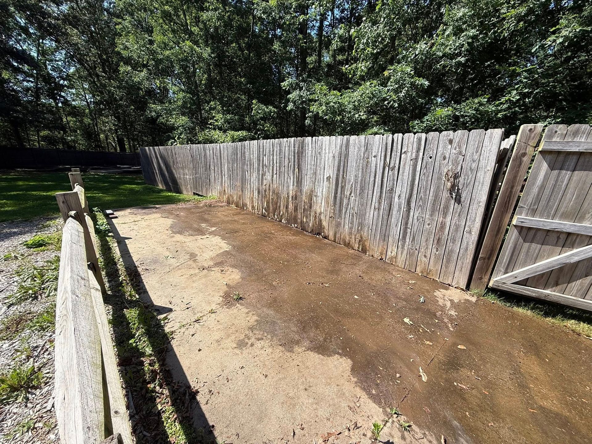 A weathered wooden fence encloses a dirt area. Green grass and trees are in the background on a sunny day.