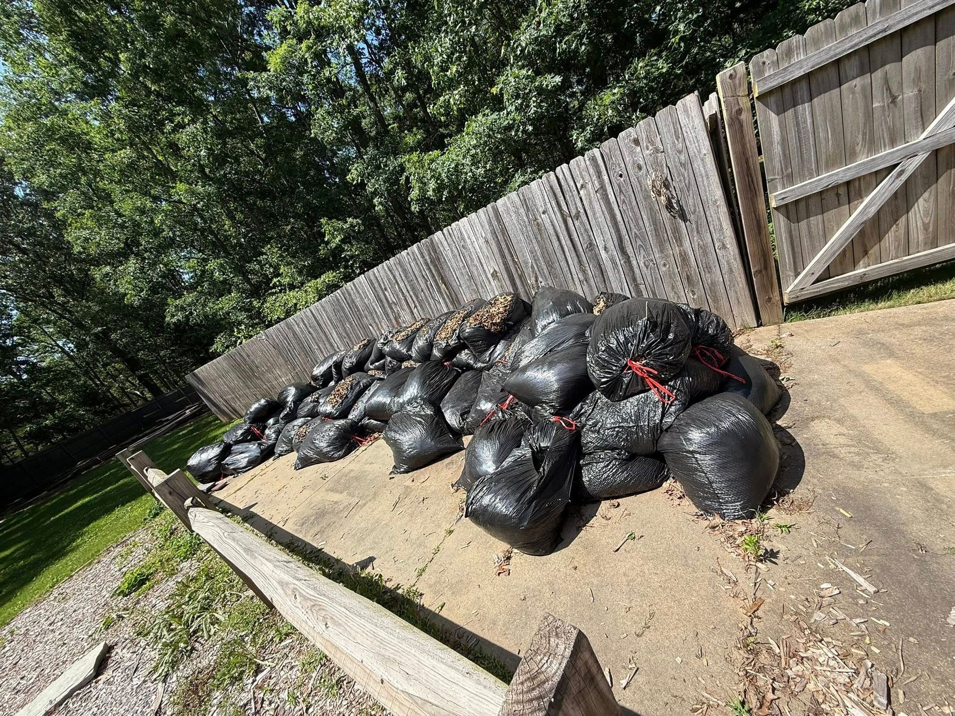 Pile of black trash bags on a concrete slab next to a wooden fence; sunny outdoor setting.