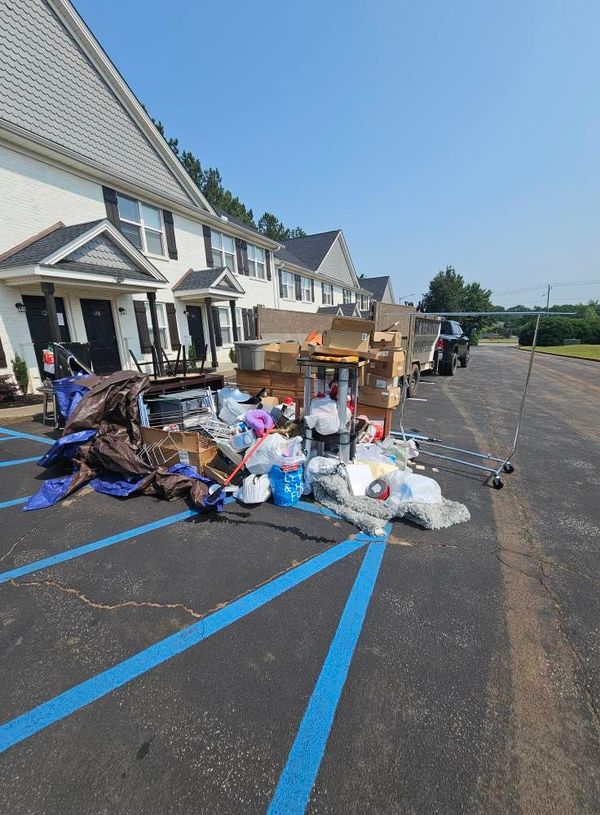 Pile of trash, boxes, and belongings in a parking lot near apartment buildings; sunny day.