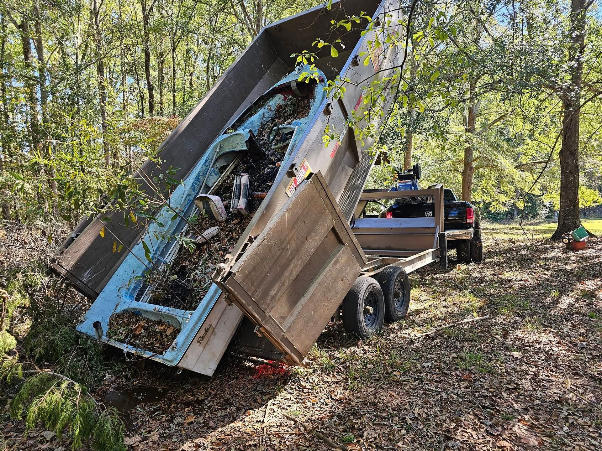 A blue dump trailer tilted, unloading debris in a wooded area; sunlight shines through the trees.