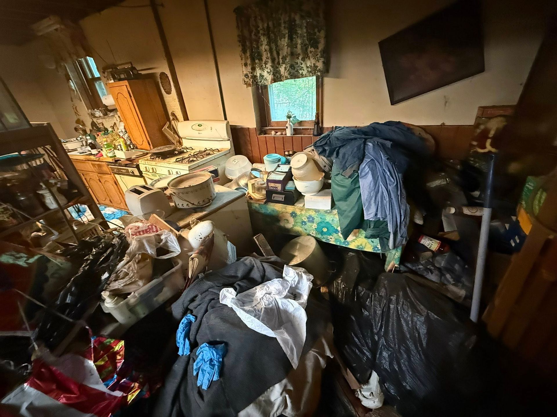 Messy, cluttered interior of a room with piles of clothing, trash, and kitchen items.