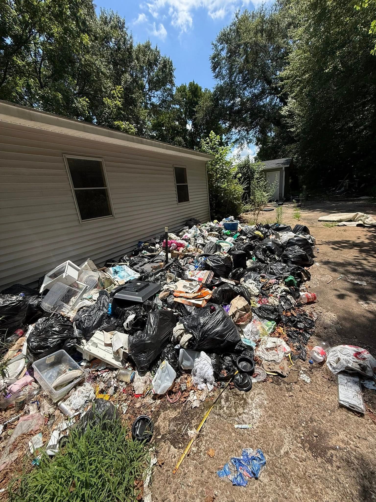 Pile of trash in a yard next to a building. The sky is blue and trees are in the background.