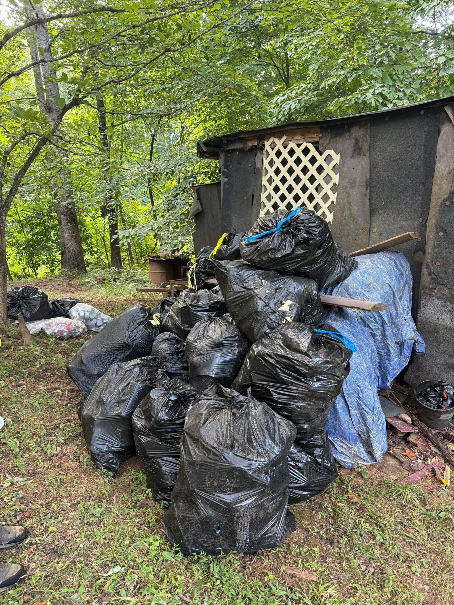 Pile of black trash bags in front of a small, dilapidated building outdoors, surrounded by trees and grass.