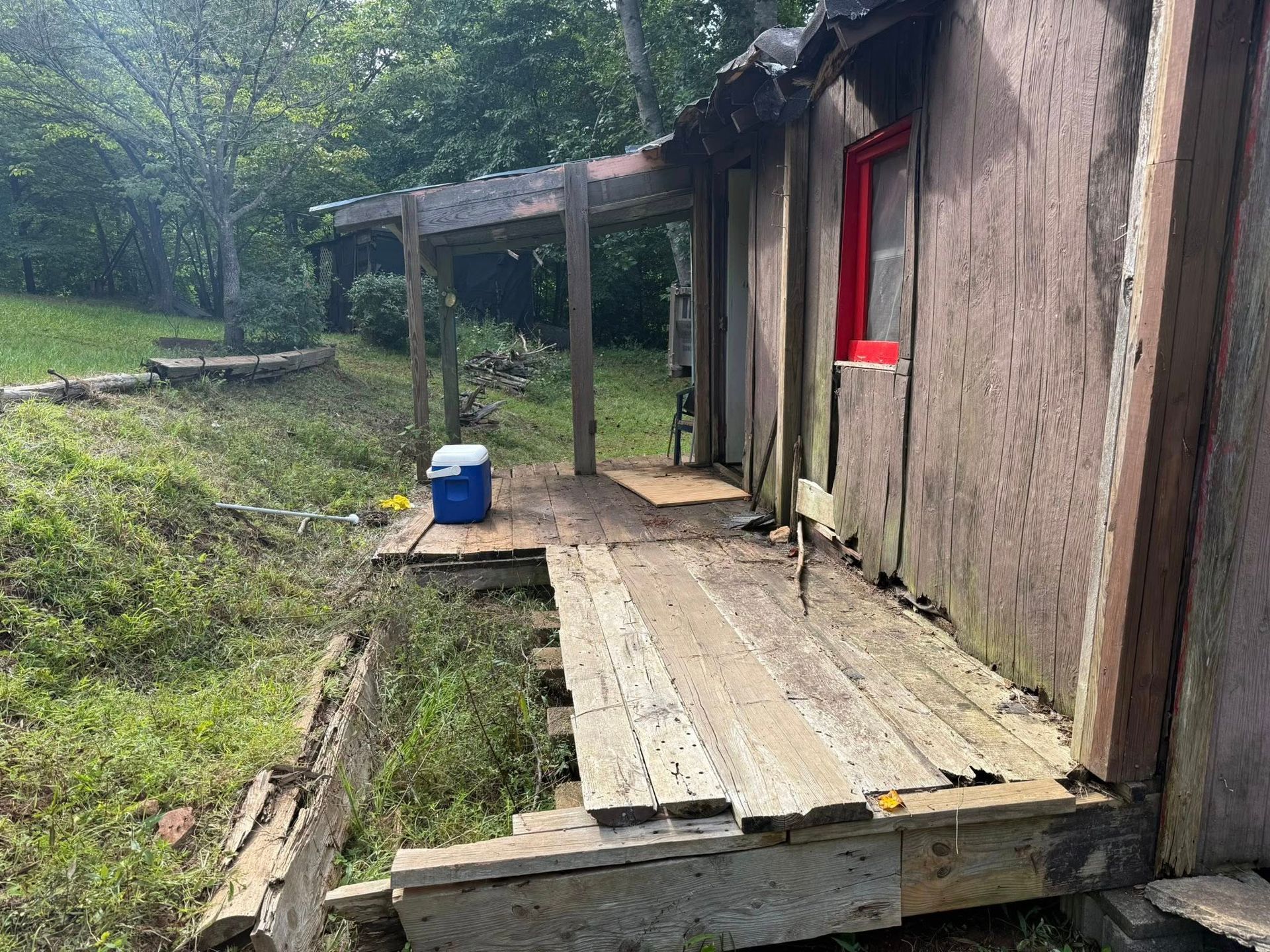 Dilapidated wooden porch with a red-framed window, surrounded by overgrown grass and trees.
