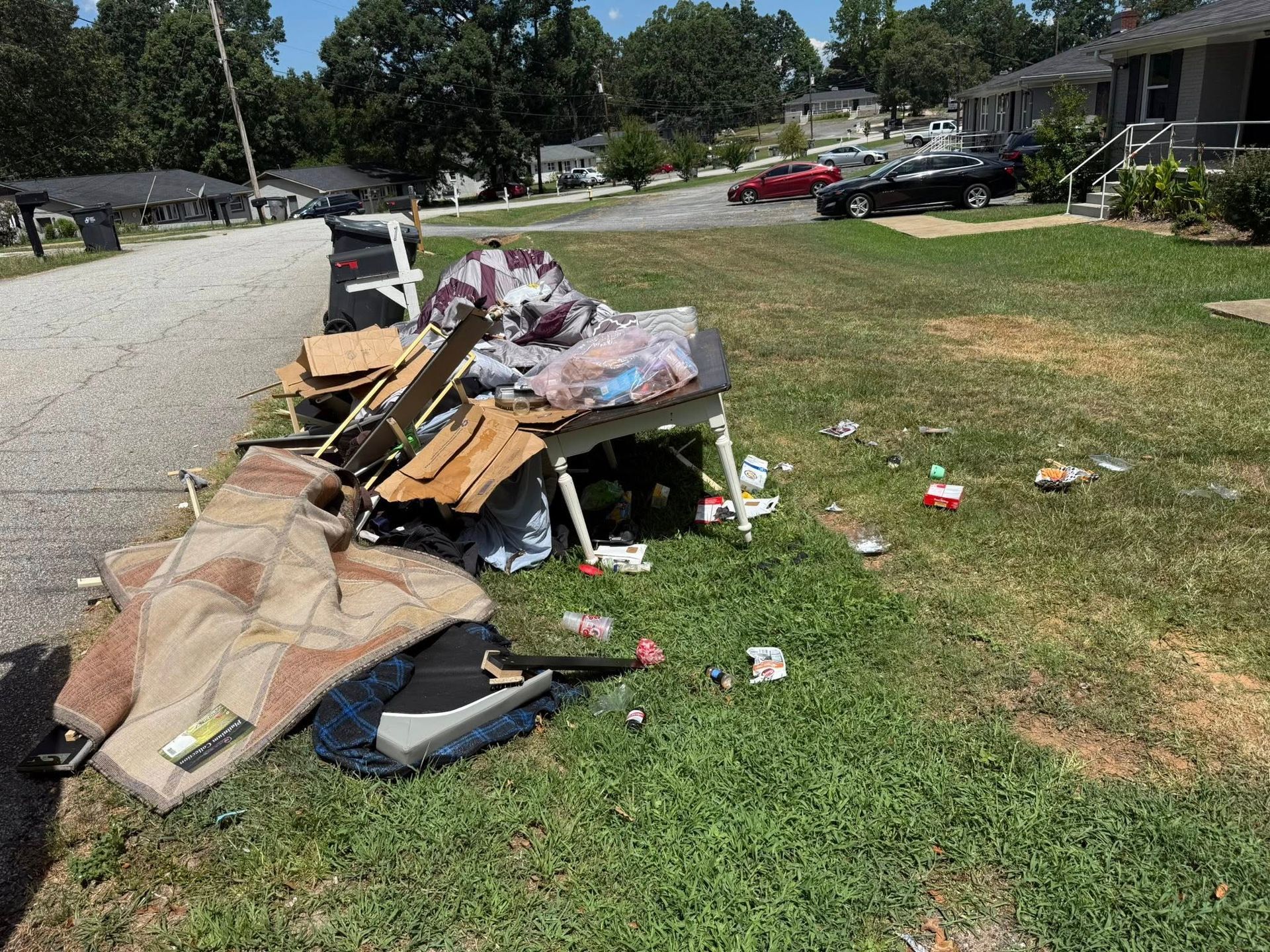 Pile of trash on a grassy lawn next to a street. Includes furniture, carpet, and scattered debris.
