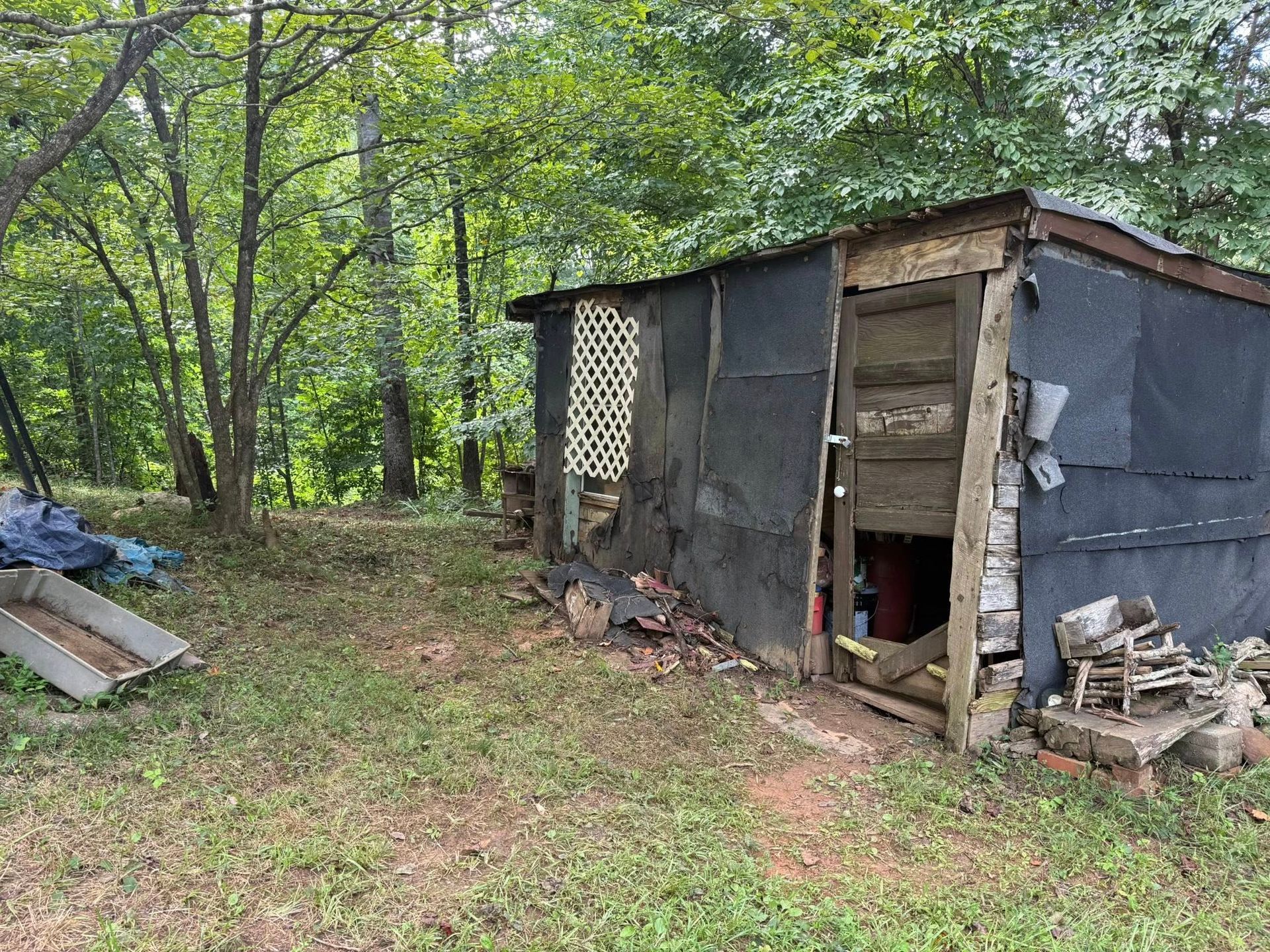 Dilapidated shed in a grassy yard, black covering, white lattice, trees in the background.