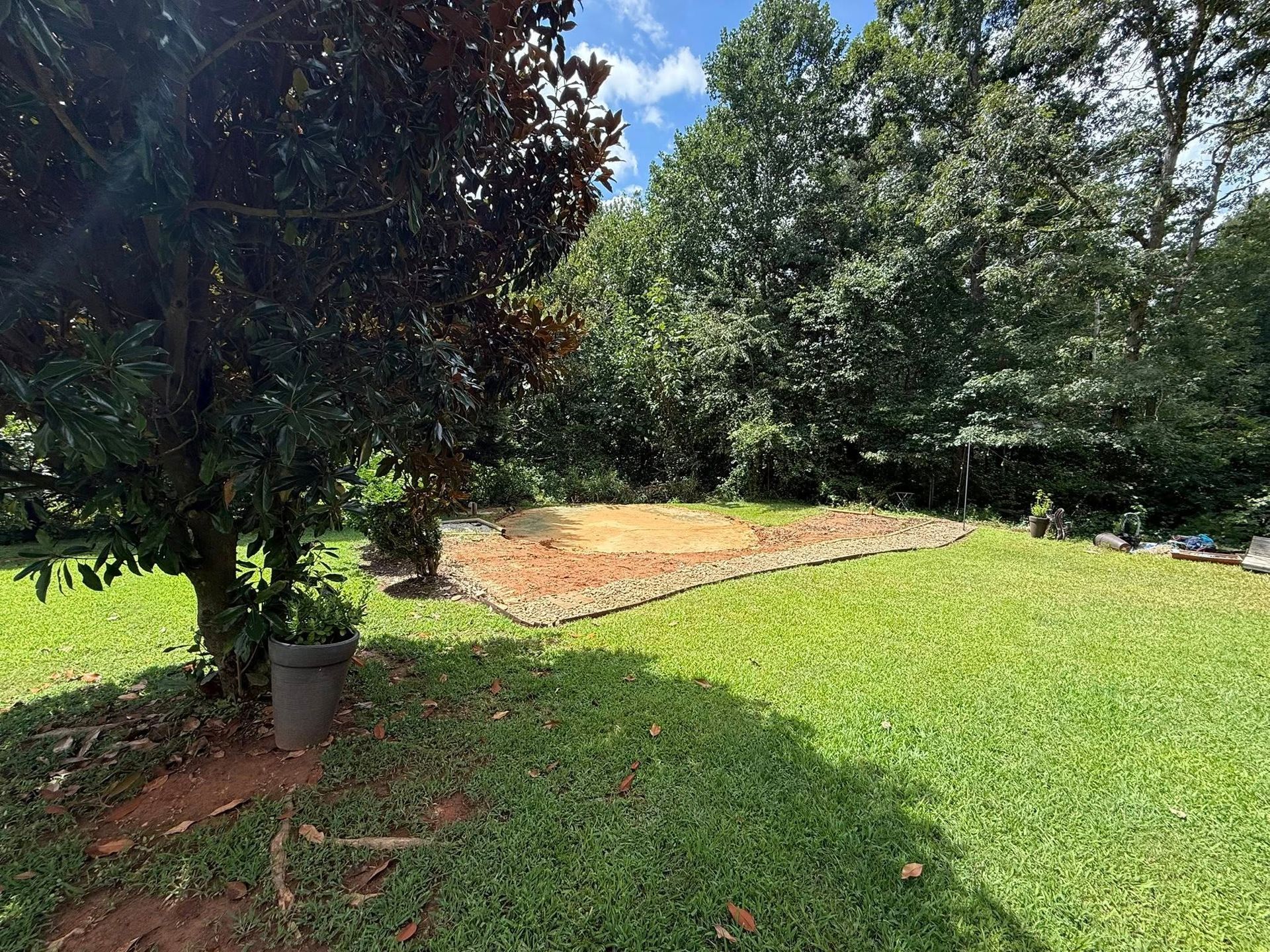 Grassy backyard with a bare patch bordered by stone, surrounded by trees. A tree on the left casts a shadow.