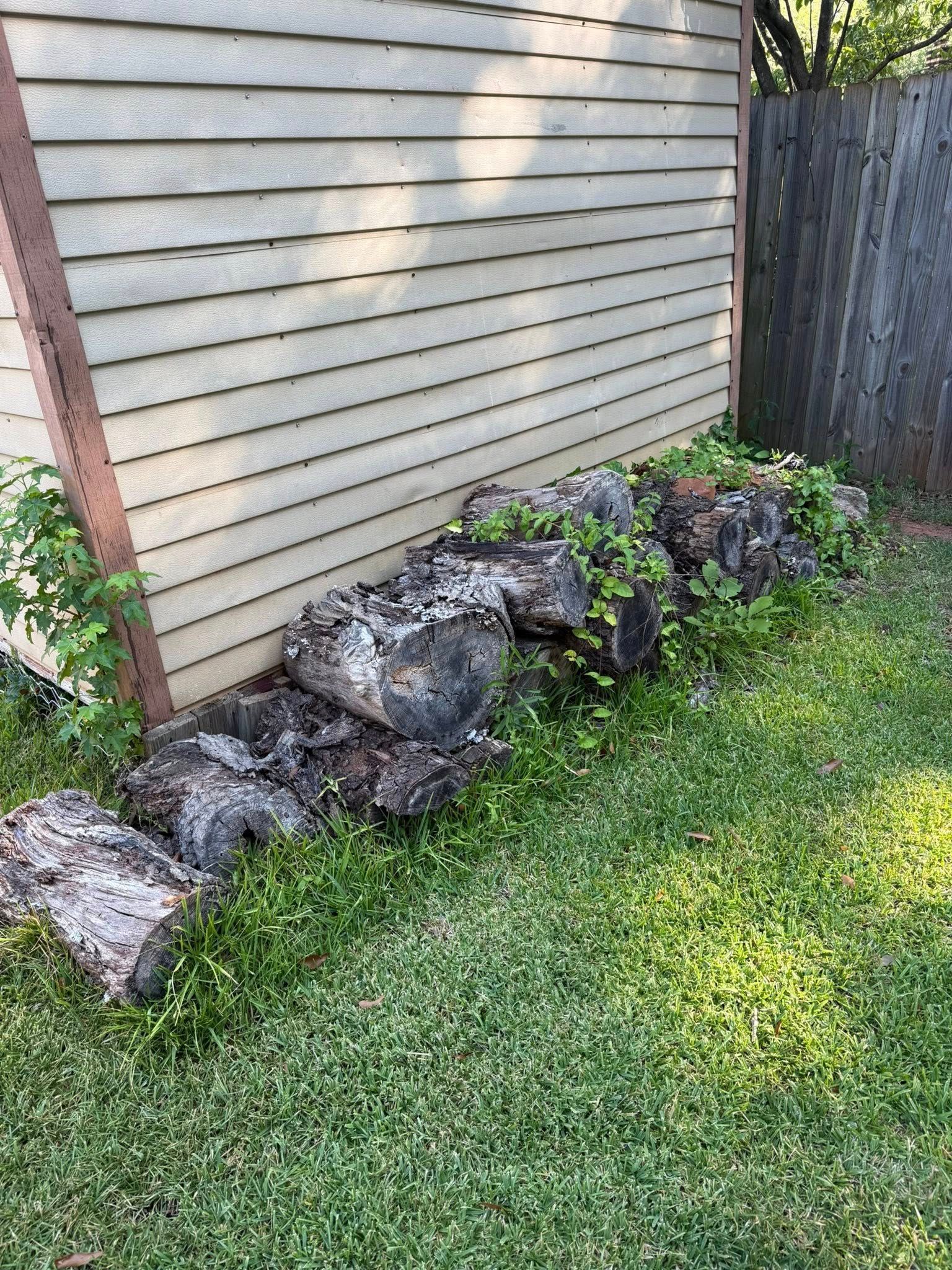 Logs stacked near a building's siding, with grass in front and a fence to the right.