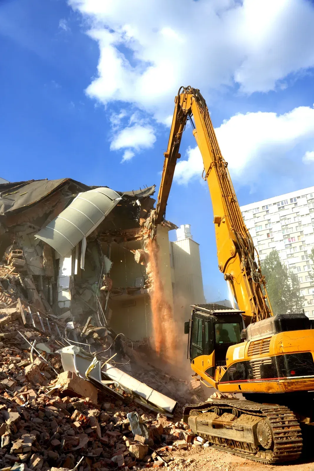 Yellow excavator demolishing a building, debris and dust in the air, blue sky in the background.