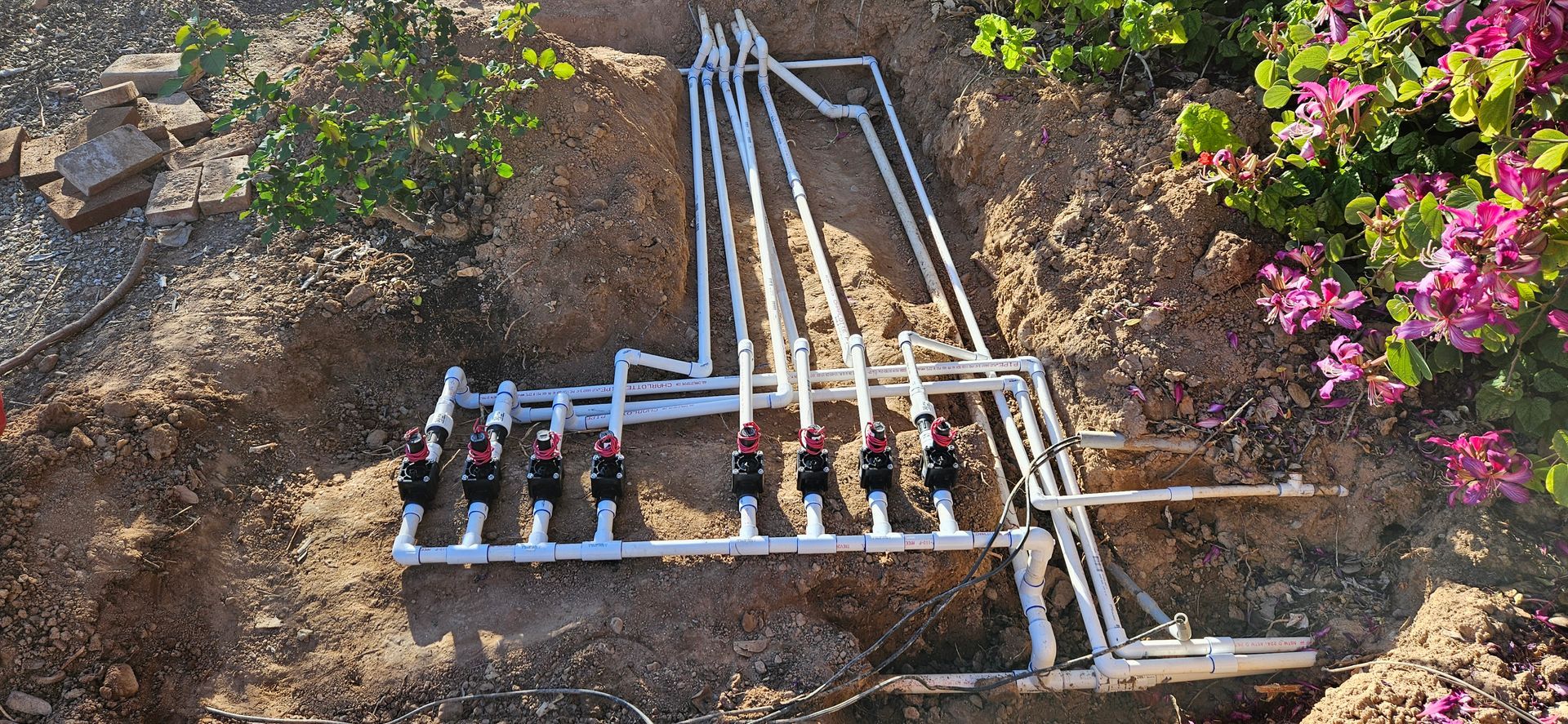 A manifold of white PVC pipes and irrigation valves installed in an open dirt trench near garden plants.