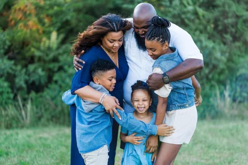 Family embraces outdoors; a woman in navy, children in blue, man in white, smiles.