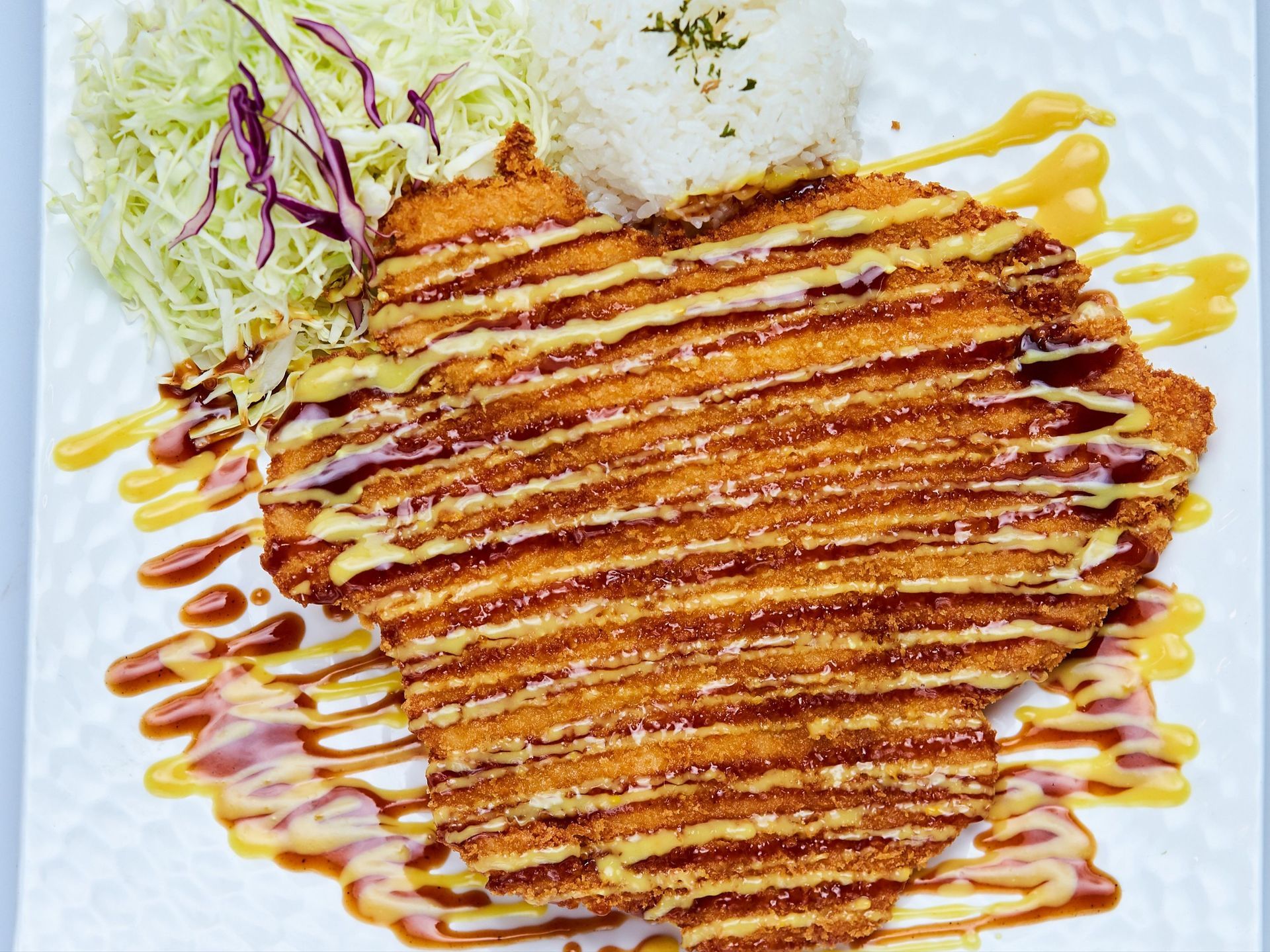 Breaded and fried chicken cutlet with rice and salad on a white plate, drizzled with sauce.