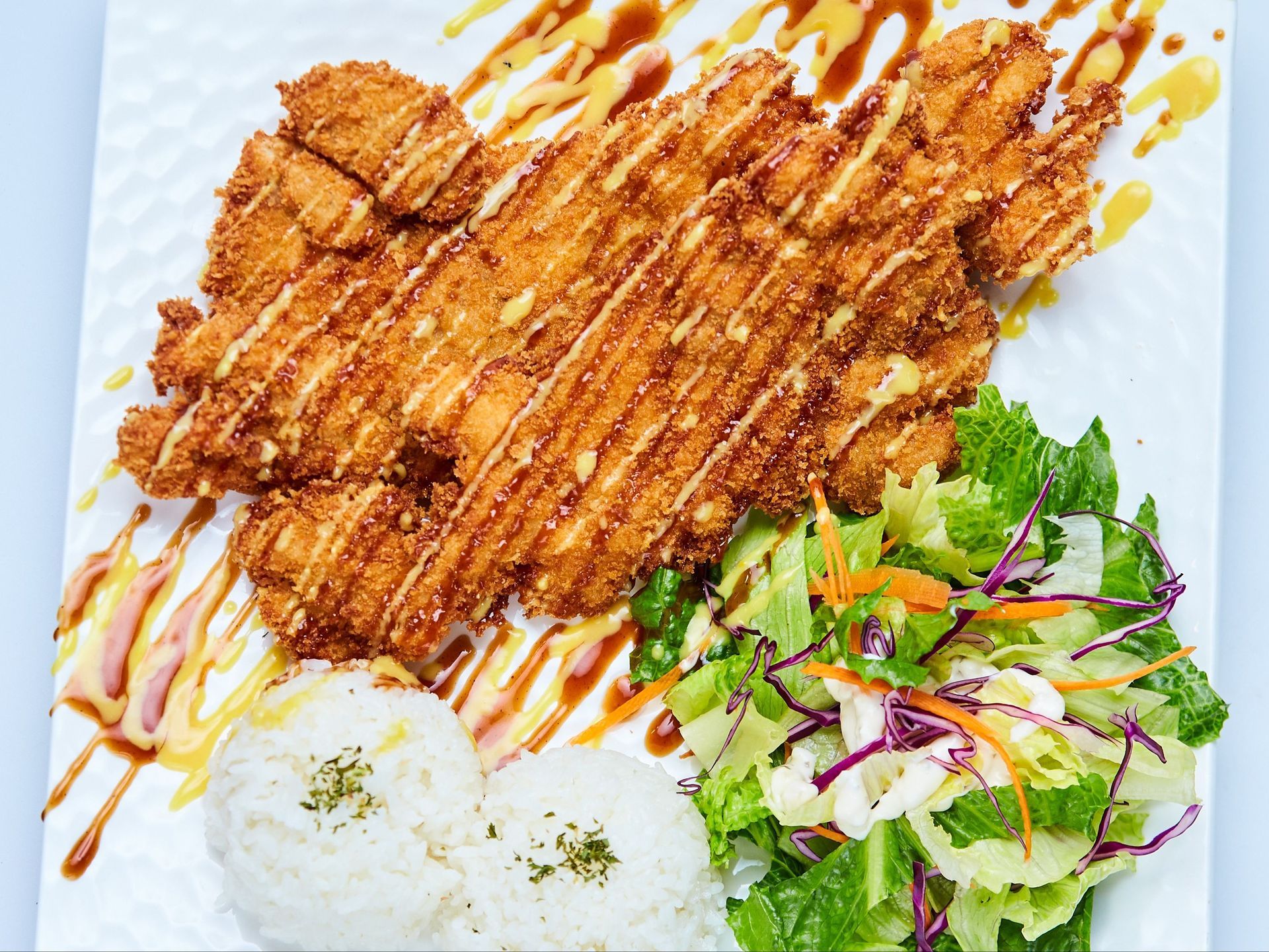 Chicken katsu with rice, salad, and drizzled sauce on a white plate.