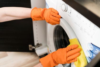Manos con guantes naranjas limpiando una lavadora de carga frontal con un paño amarillo.