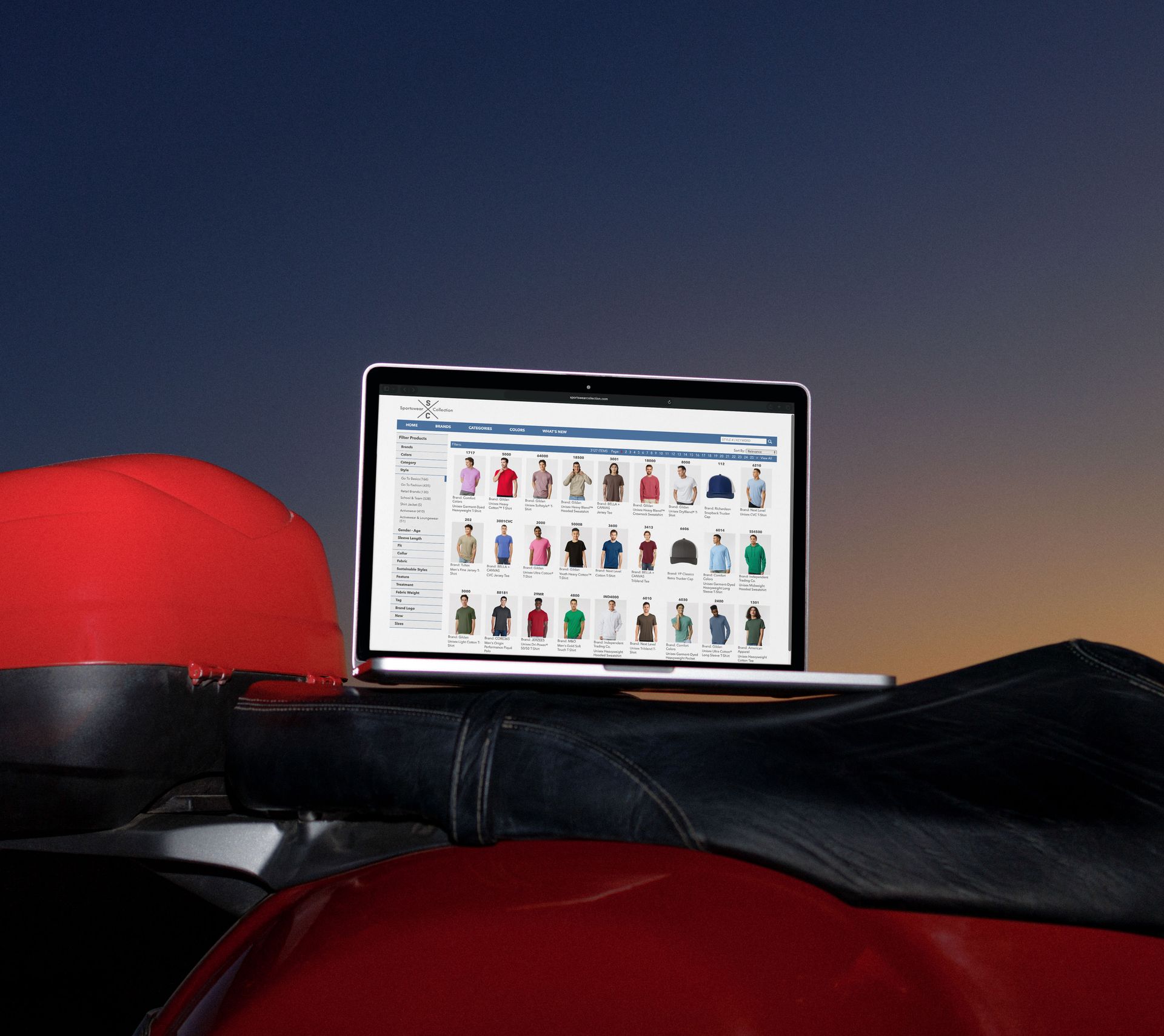 Laptop displaying clothing options sits on a red motorcycle seat. Blue sky in background.