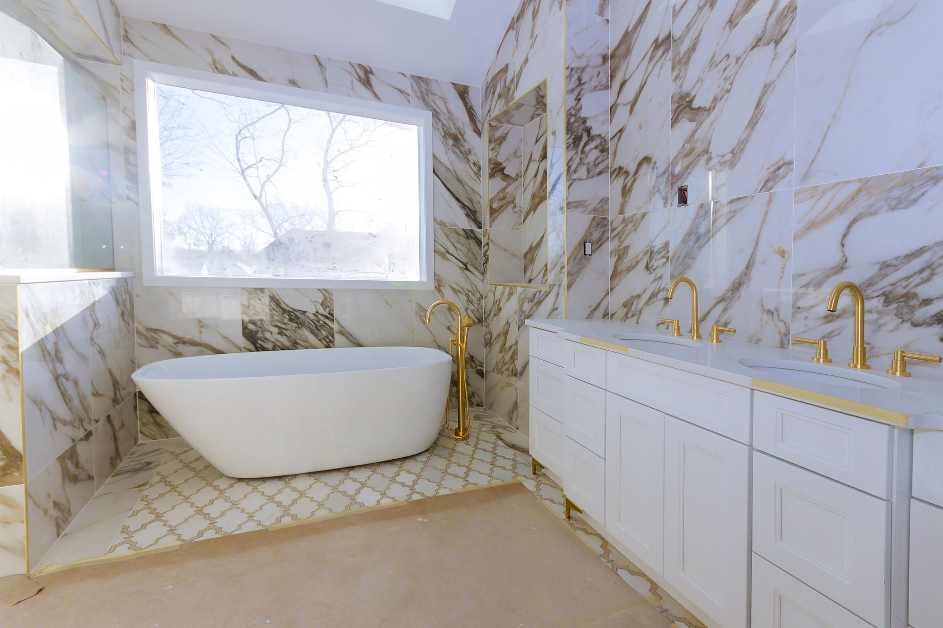 Bright marble bathroom with a freestanding tub, gold fixtures, and a white vanity beneath a large window