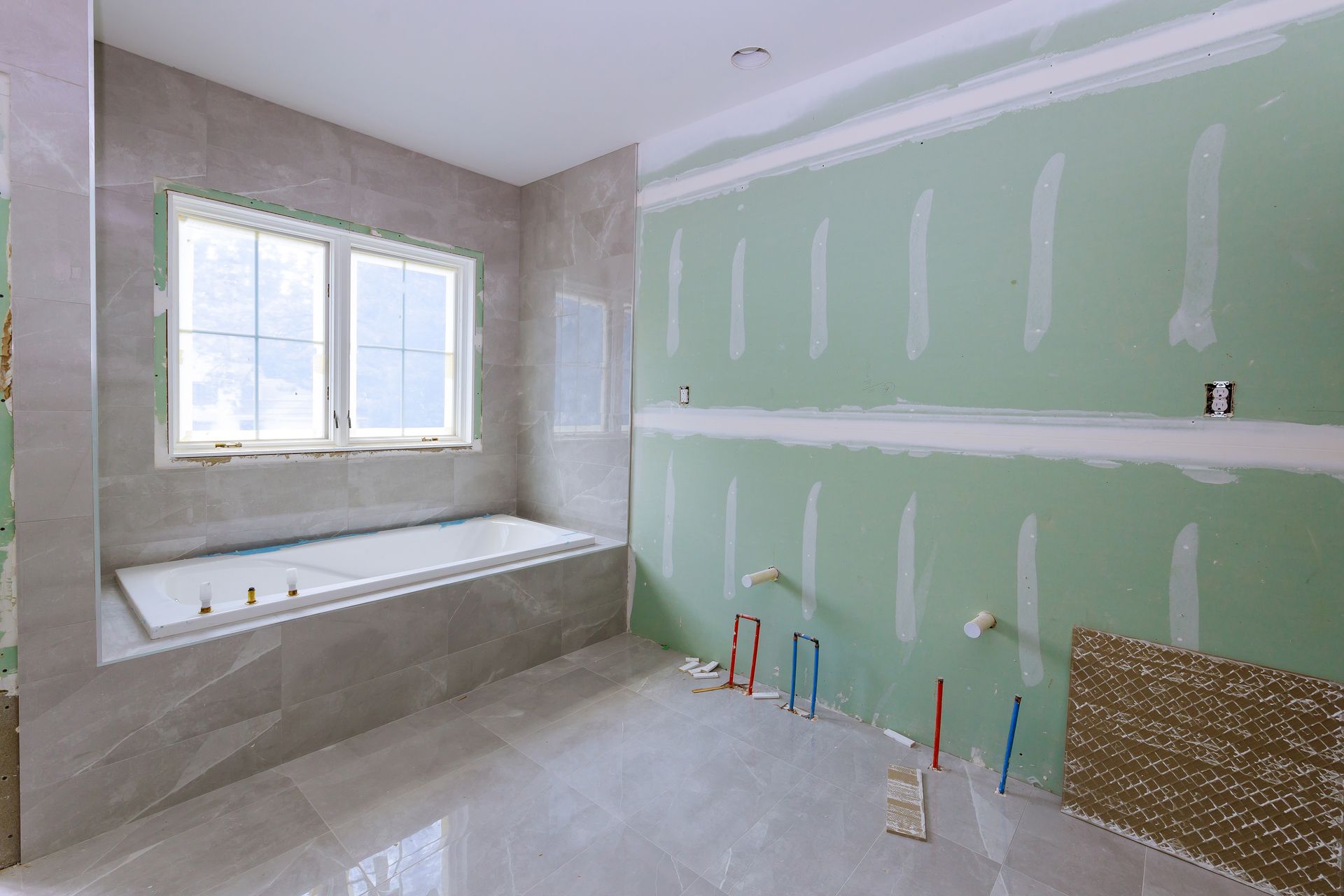 Bathroom under renovation with green drywall, tub by window, and unfinished floor.